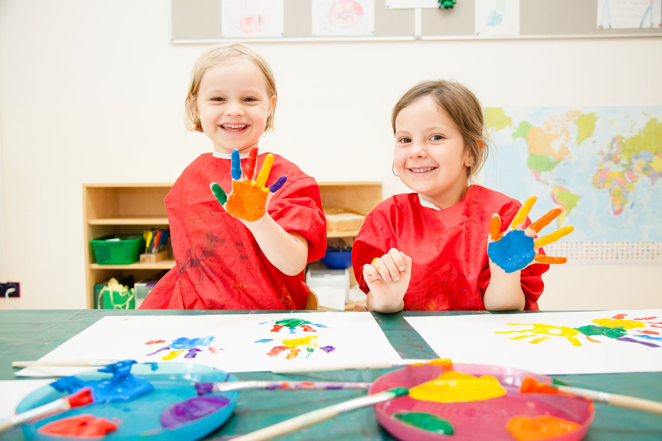 Zwei Mädchen experimentieren im Kindergarten mit Fingerfarben und halten ihre bunt bemalten Hände in die Kamera