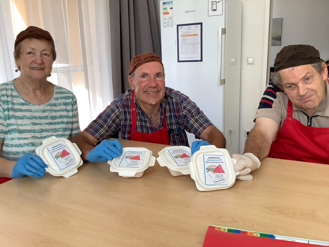 Zwei Männer und eine Frau sitzen an einem Tisch und halten nachhaltige Jausendosen in die Kamera. Sie tragen blaue Handschuhe, Kopfbedeckungen und rote Schürzen.