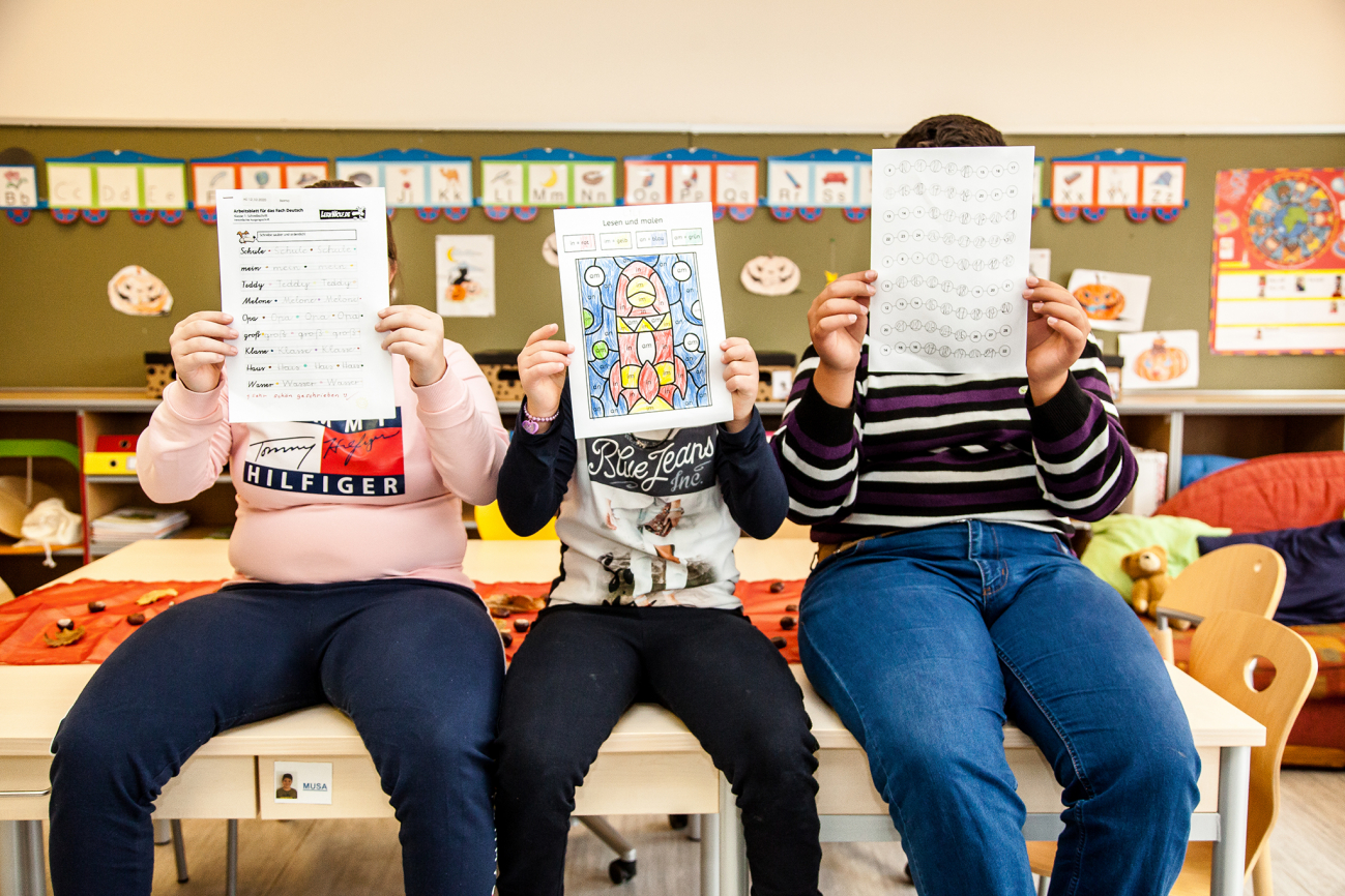 Drei Schulkinder sitzen auf einem Tisch und halten ihre Übungsblätter vor ihre Gesichter.