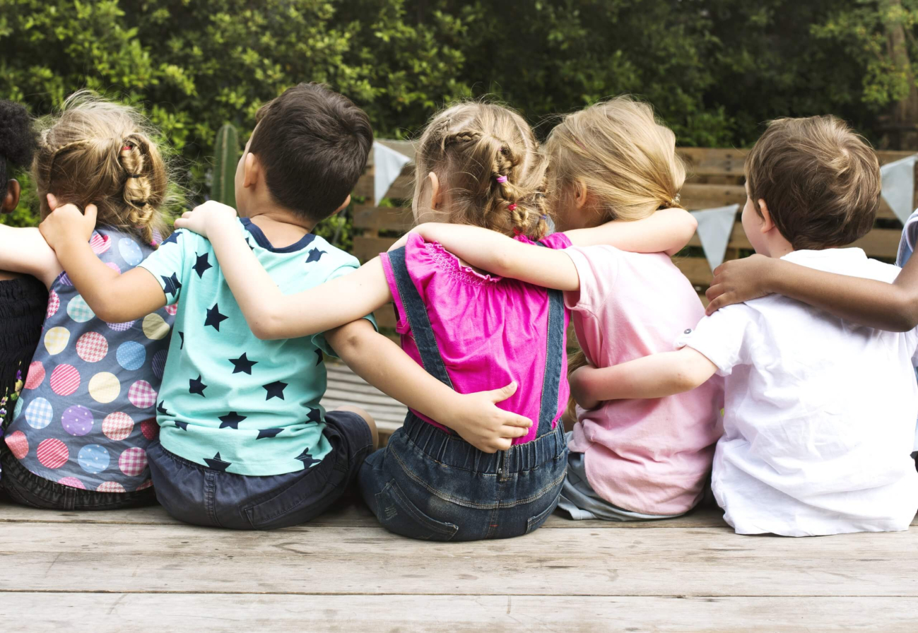 Eine Gruppe von Kindergartenkindern von hinten auf einer Bank im Freien. Die Kinder halten einander an den Schultern.