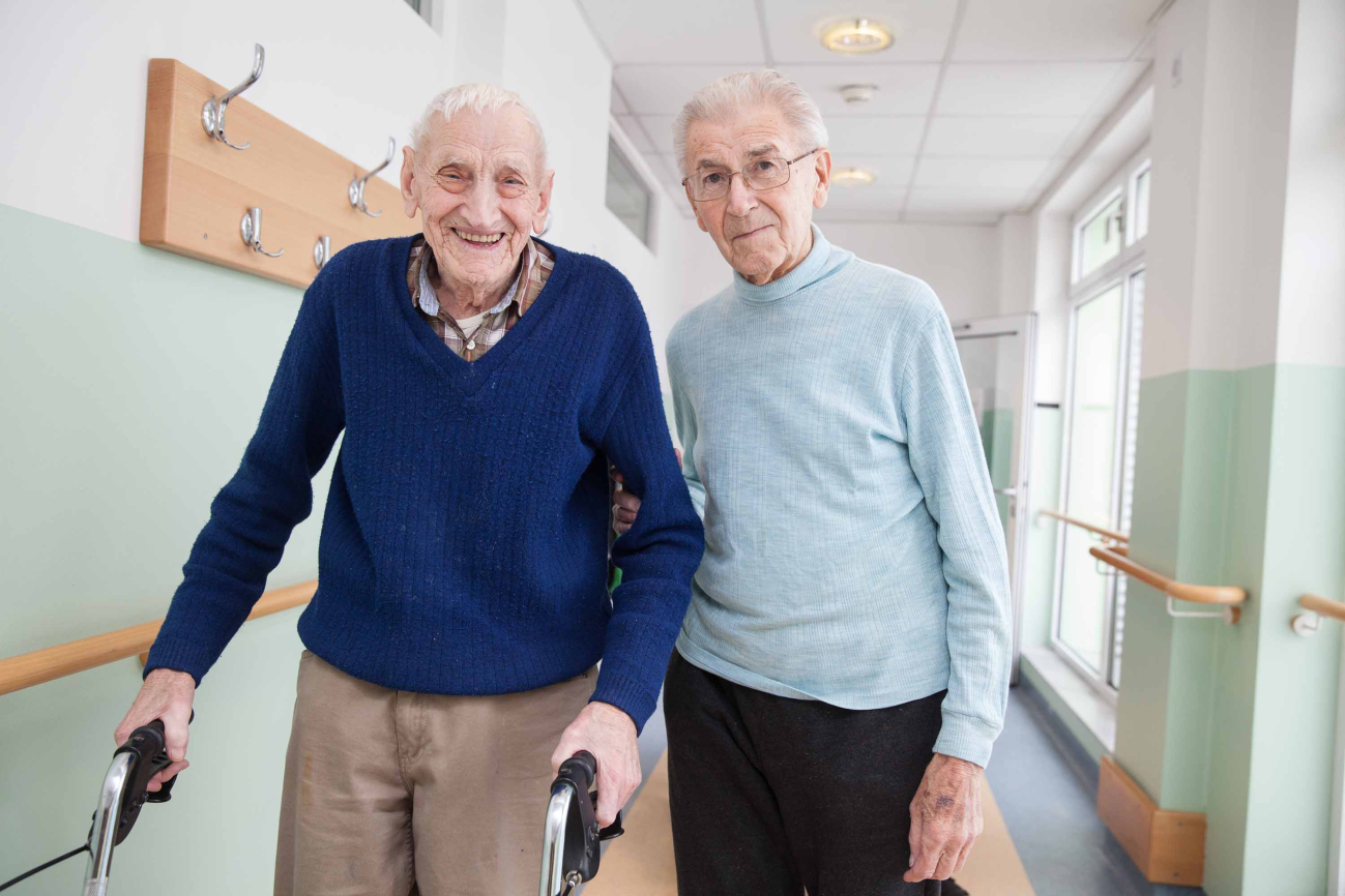 Ein Mann im Rollator wird von einem angehörigen begleitet.