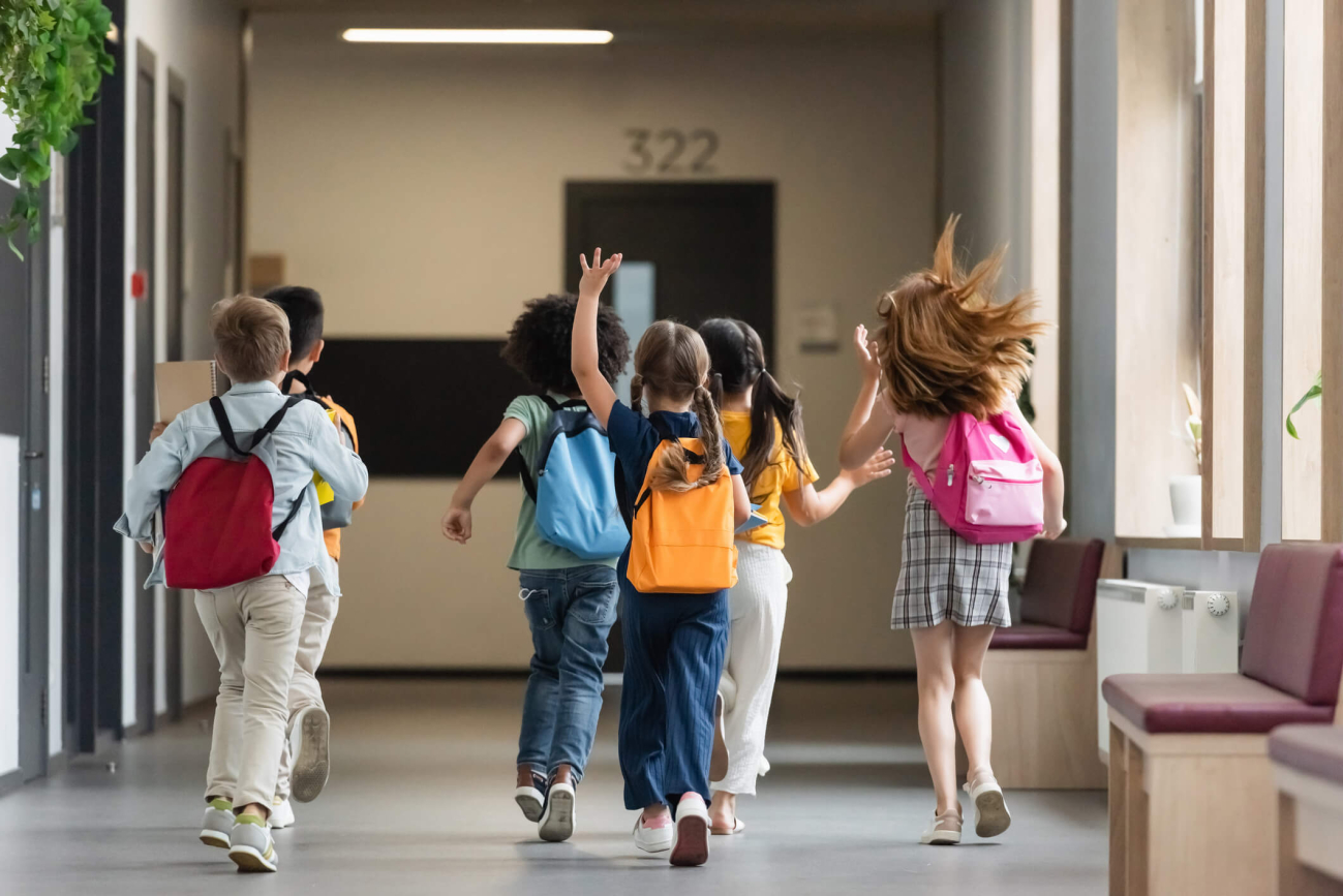 Mehrere Volksschulkinder laufen in Richtung ihres Klassenzimmers. Ein Mädchen hebt fröhlich die Hand.