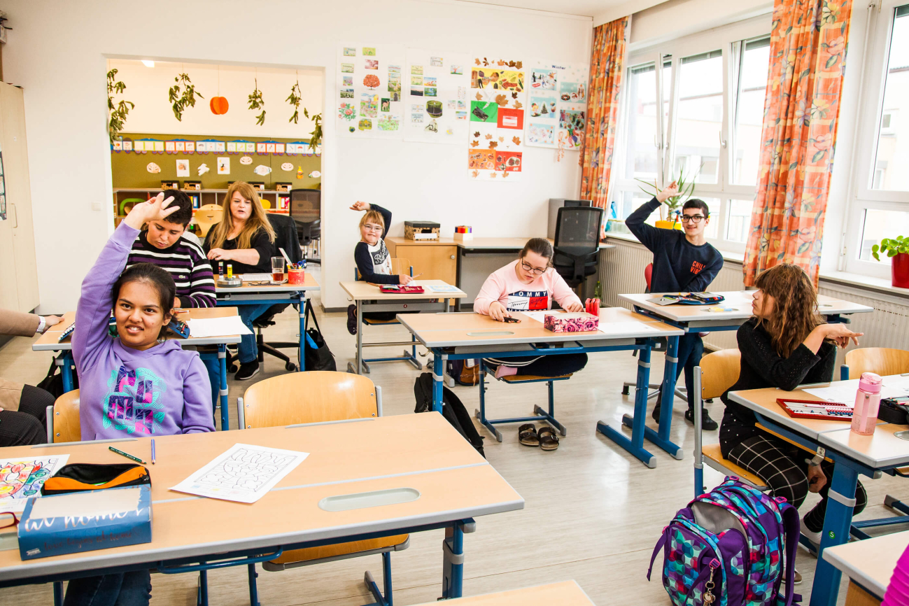 Eine Schulkasse mit Schüler:innen. EInige der Schüler:innen zeigen auf. Eine der Schüler:innen ist Hoffnungsträgerin Lili.