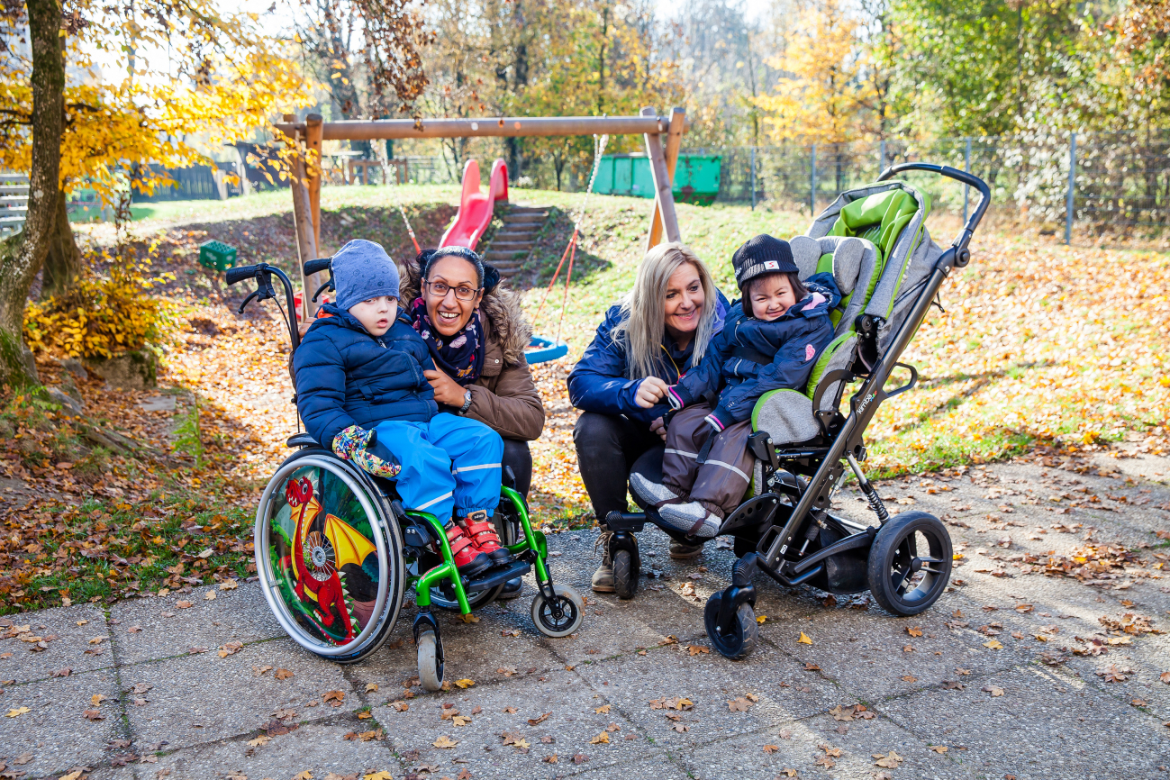 Junge im Rollstuhl und Mädchen im Buggy im Garten, begleitet von Elementarpädagog:innen