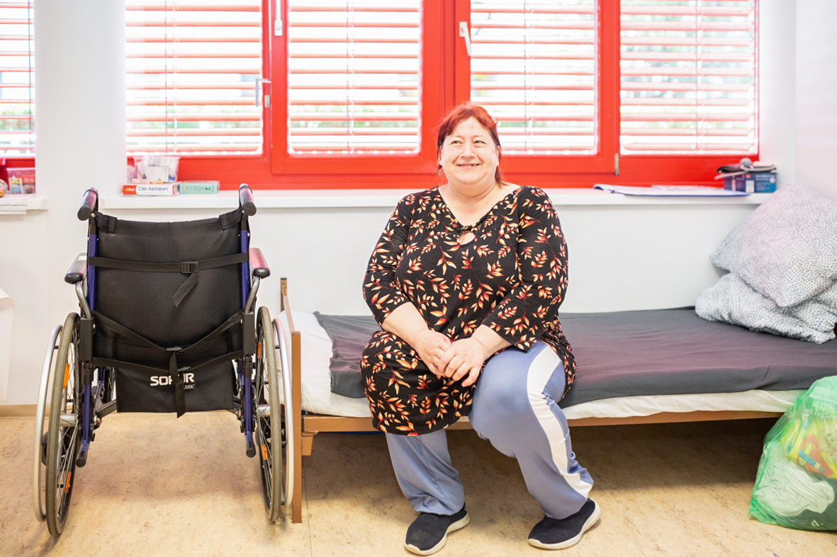 Frau sitzt lächelnd auf ihrem Bett, daneben steht ein Rollstuhl