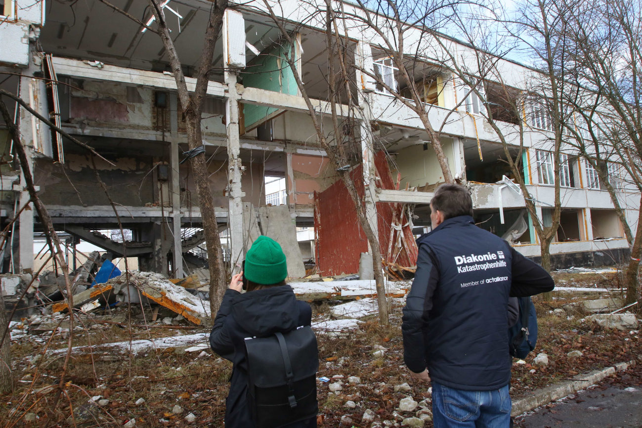 Mitarbeiter der Diakonie Katastrophenhilfe stehen vor einem völlig zerbombten Schulgebäude.