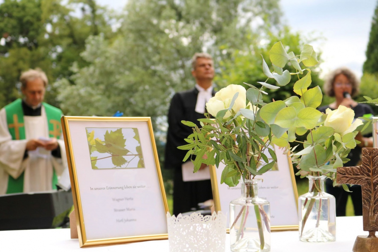 Blumenschmuck im Außenbereich des Hauses am Ruckerlberg, Bilderrahmen, ein Metallkreuz, im Hintergrund verschwommen drei Personen