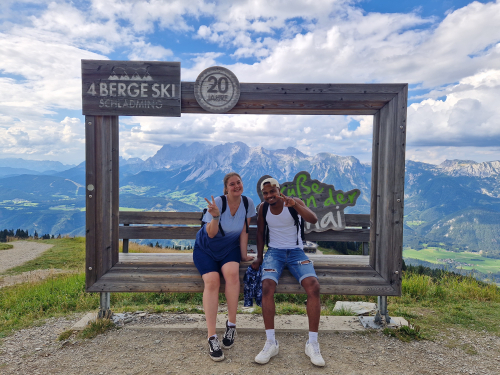 Eine junge Frau und ein junger Mann sitzen vor einem überdimensionalem Bilderrahmen im Hintergrund Bergkulisse