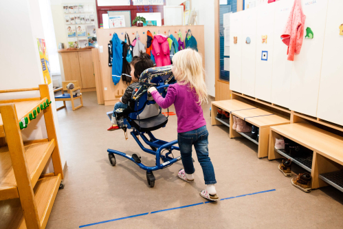 Ein Mädchen schiebt ein Mädchen im Rollstuhl durch einen Kindergarten.