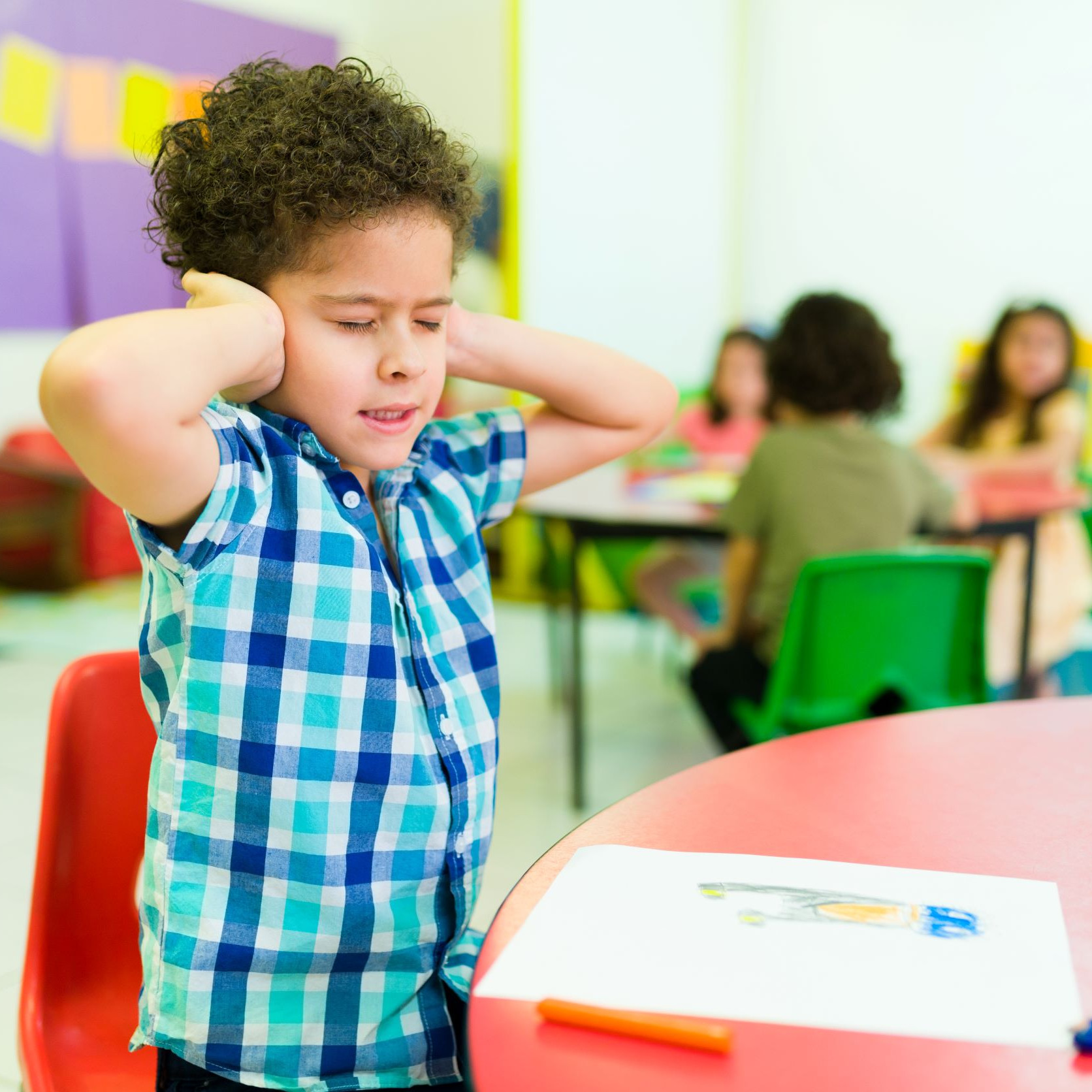 Kind steht an einem Tisch und hält sich die Ohren zu. Am Tisch hinter ihm sitzen andere Kinder.