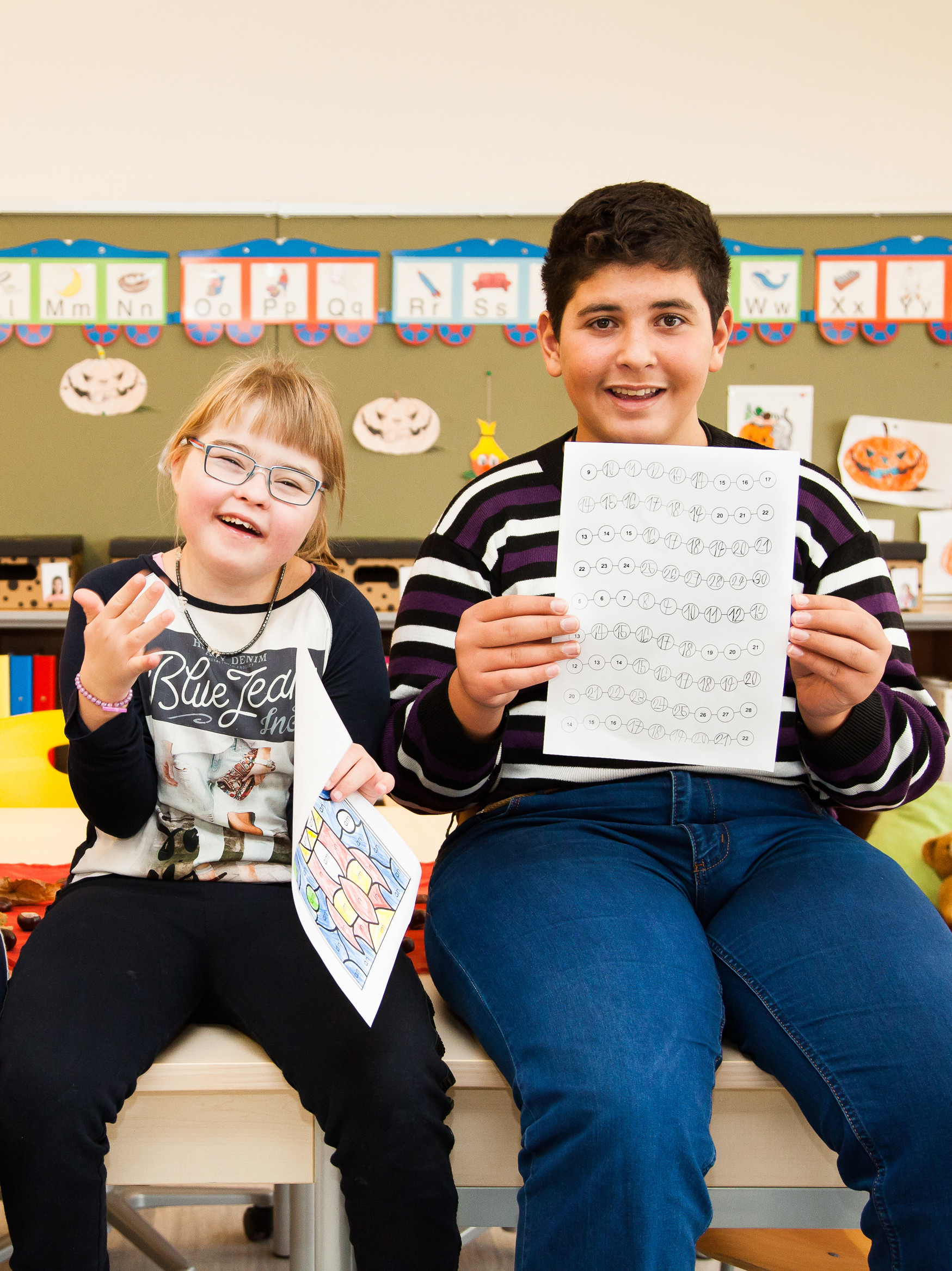 Drei Kinder mit und ohne Behinderungen lachen in die Kamera. Sie sitzen auf Tischen und halten Übungsblätter hoch.
