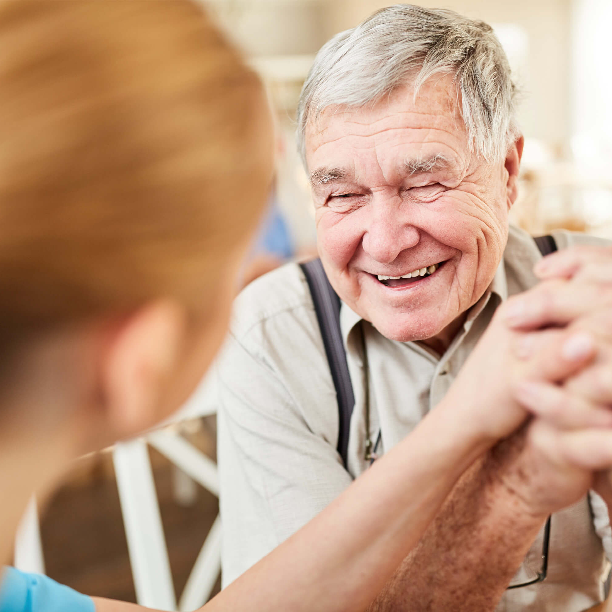 Eine Pflegemitarbeiterin und ein älterer Mann halten einander die Hände und lachen gemeinsam.
