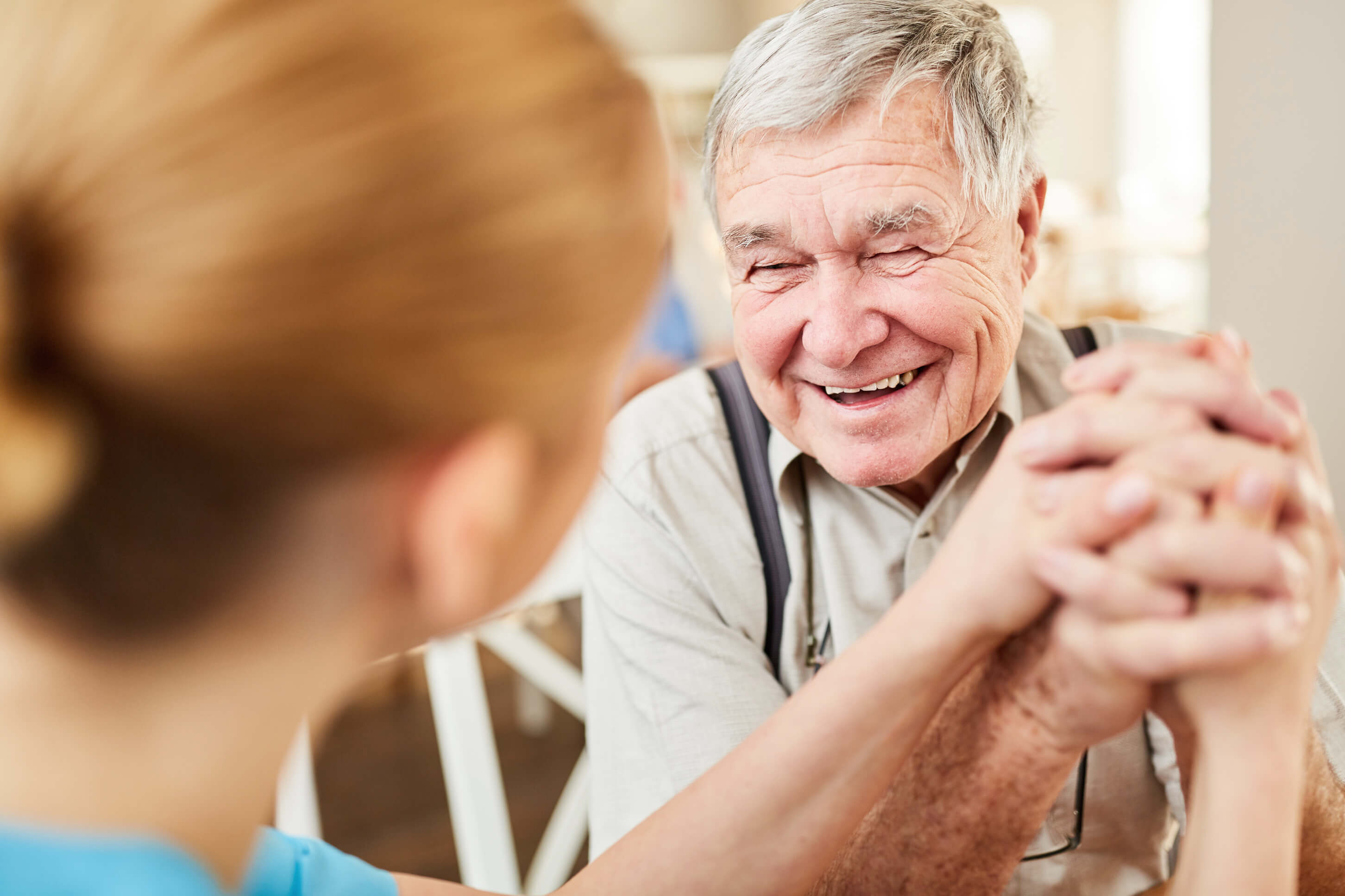 Eine Pflegemitarbeiterin und ein älterer Mann halten einander die Hände und lachen gemeinsam.