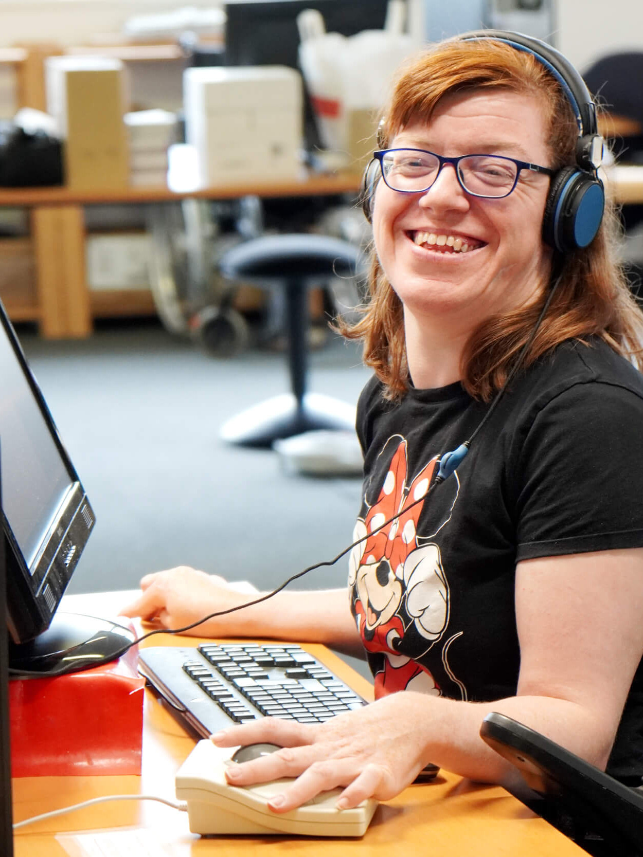 Foto einer Person im Rollstuhl die hinter einem Computer sitzt und in die Kamera lächelt
