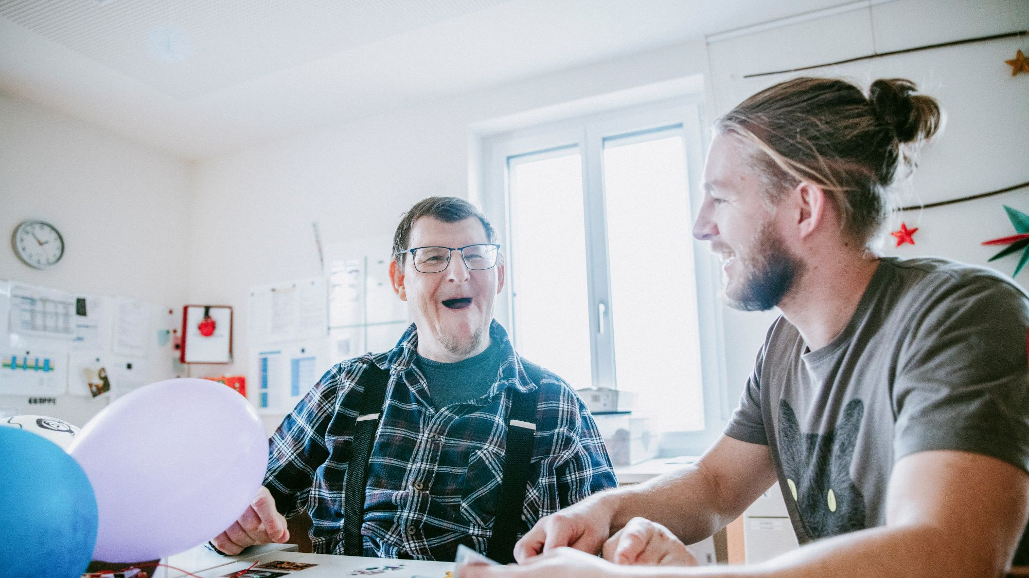 Zwei Personen sitzen nebeneinander bei Tisch, vor ihnen zwei Luftballons und Puzzlesteine