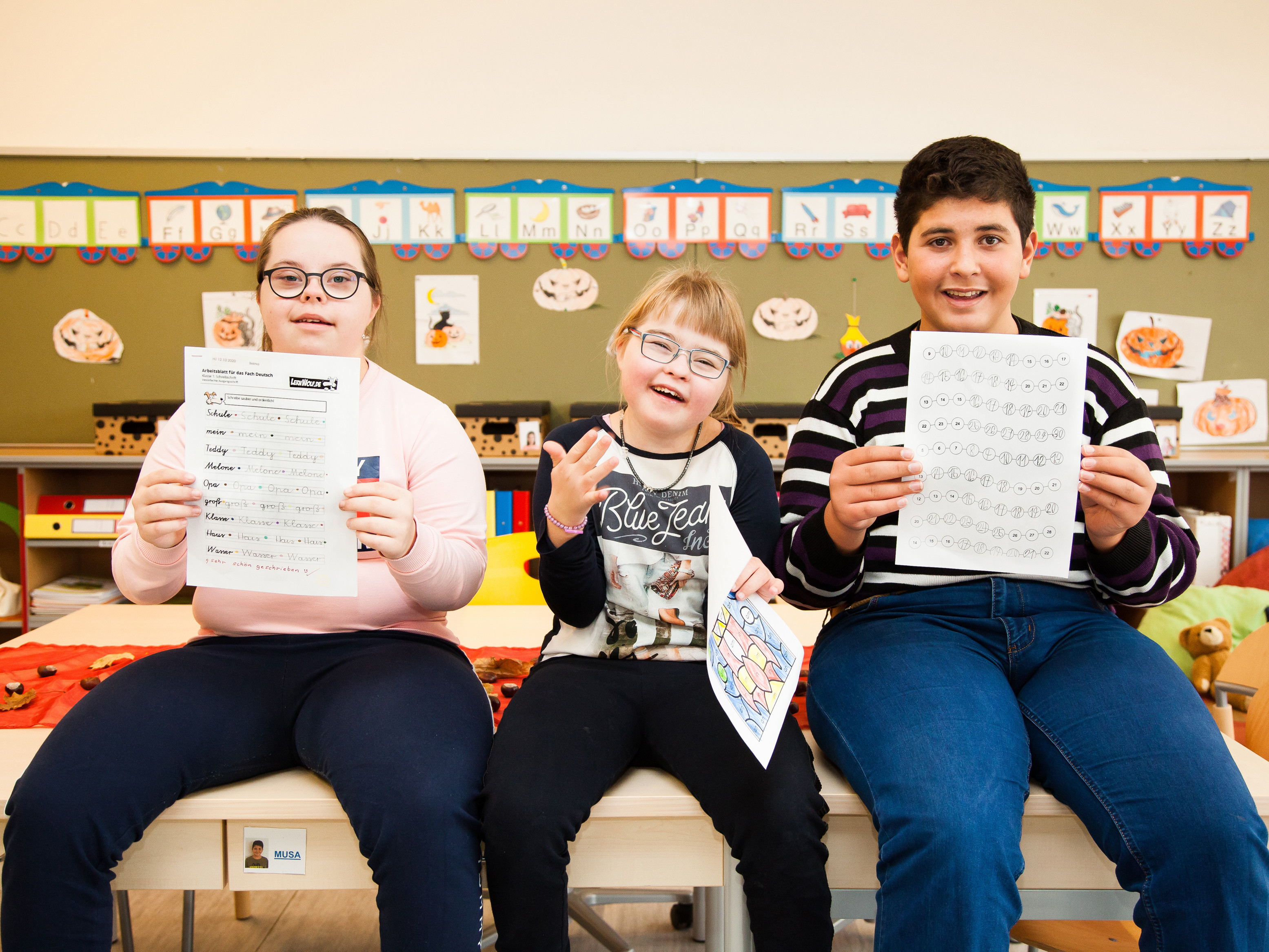 Drei Kinder mit und ohne Behinderungen lachen in die Kamera. Sie sitzen auf Tischen und halten Übungsblätter hoch.