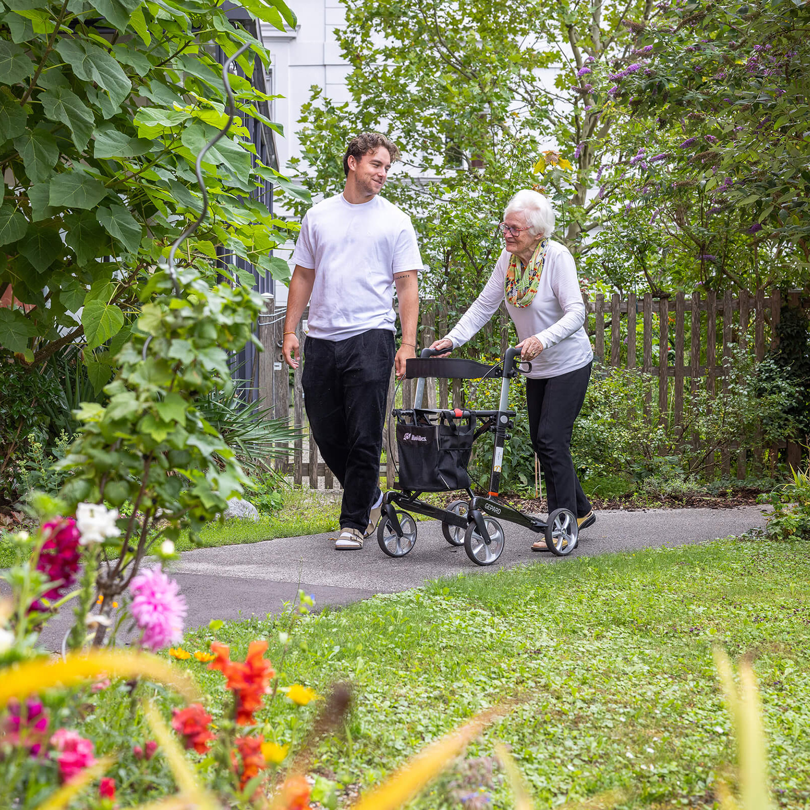 Ein Person mit Rollator und ein Pfleger unterhalten sich während eines Spazierganges im Garten
