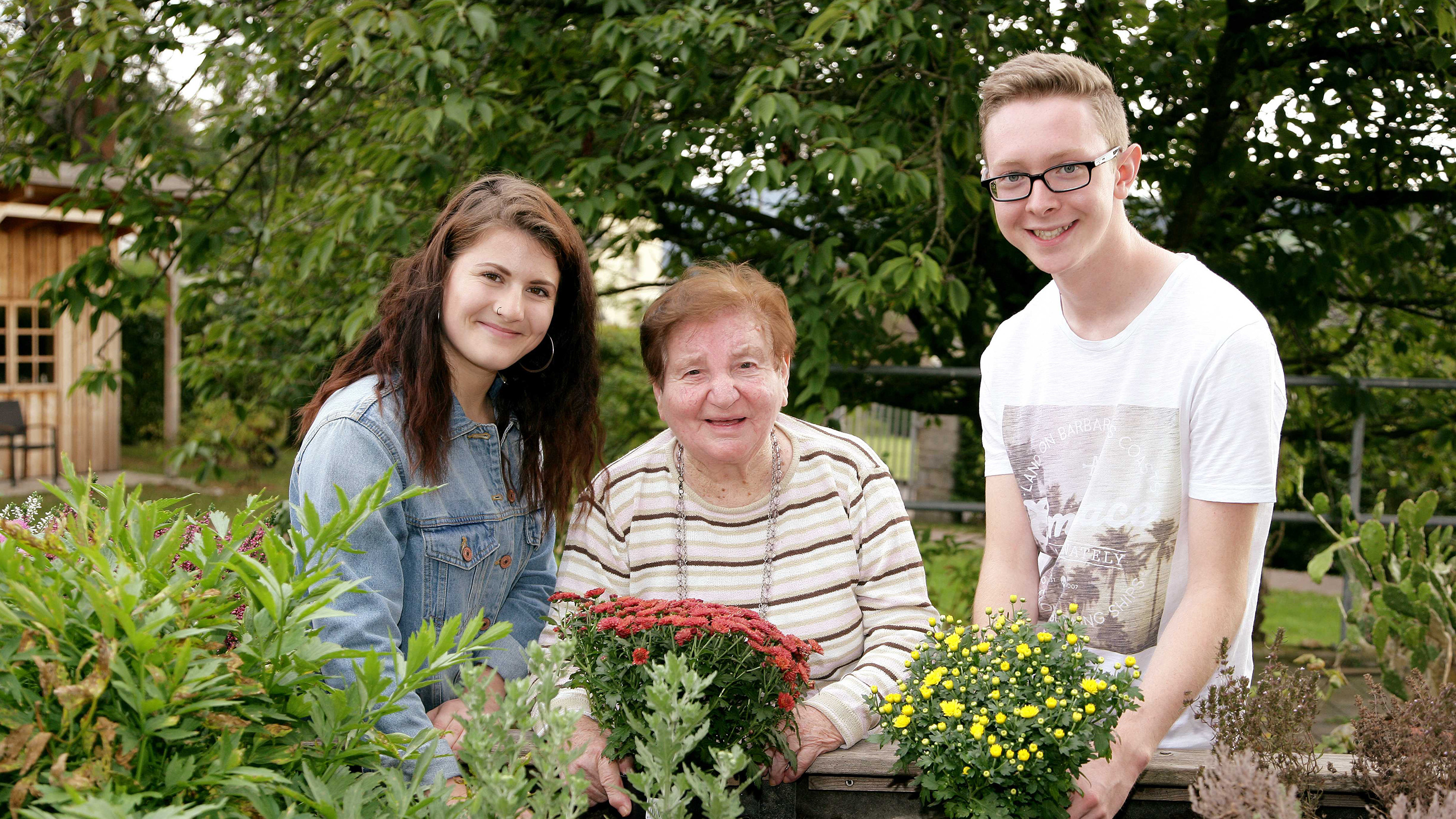 Zwei junge Menschen und eine Frau im Alter setzen gemeinsam Blumen in ein Hochbeet.