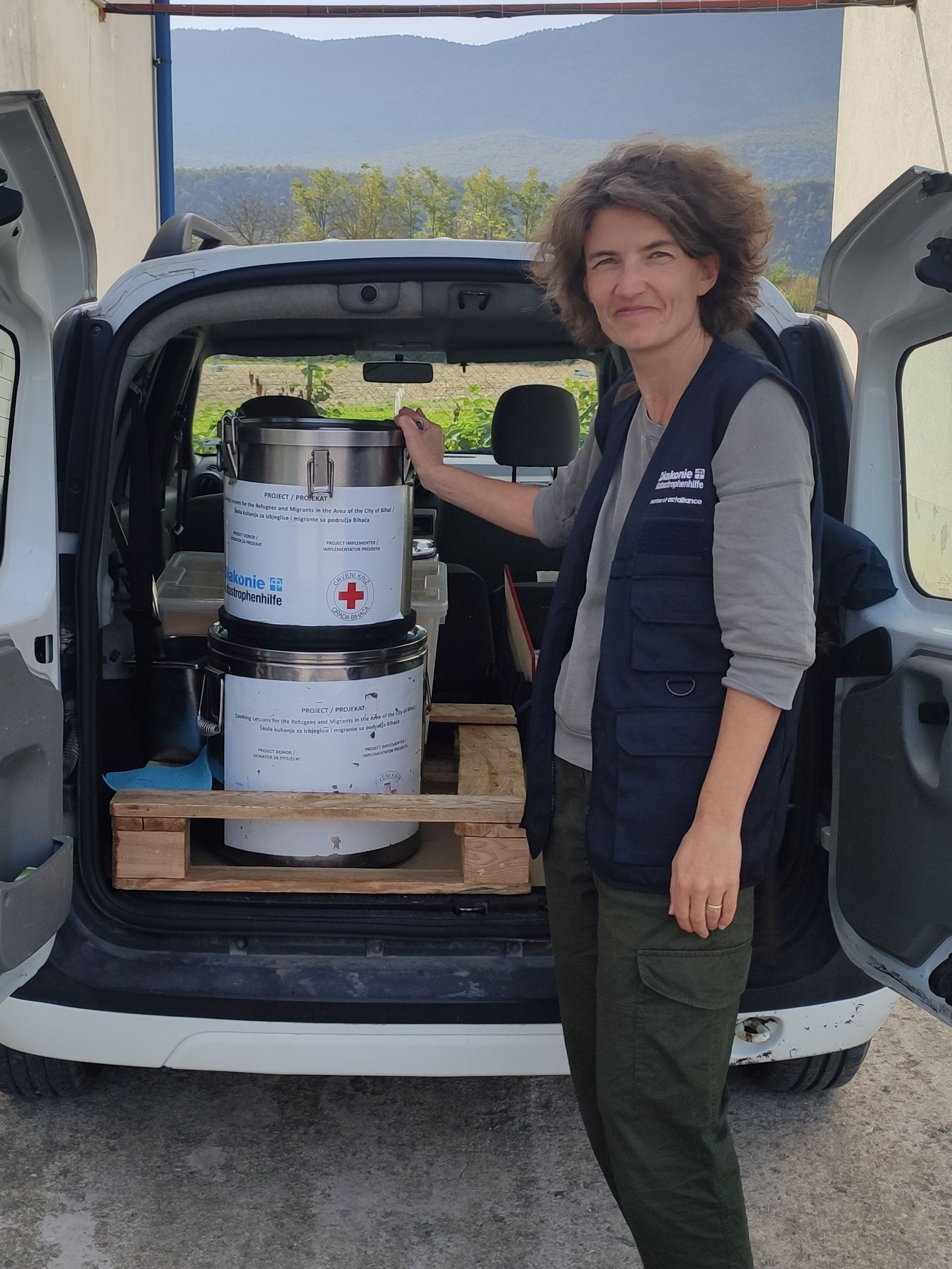 Mitarbeiterin der Diakonie Katastrophenhilfe steht vor einem Auto, das mit großen Essenstöpfen beladen ist.