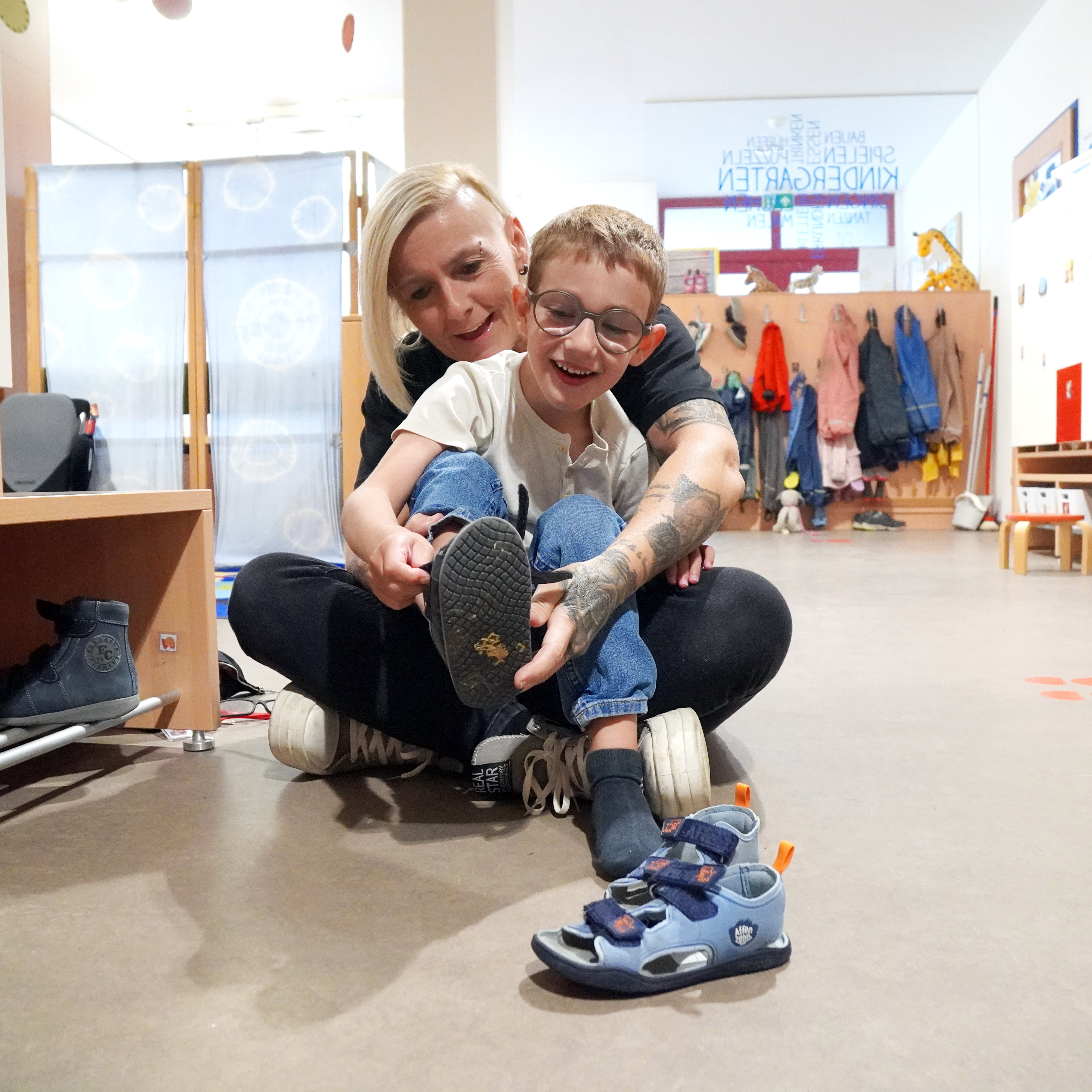Ein Kind zieht sich die Schuhe und eine Elementarpädagogin hilft dabei