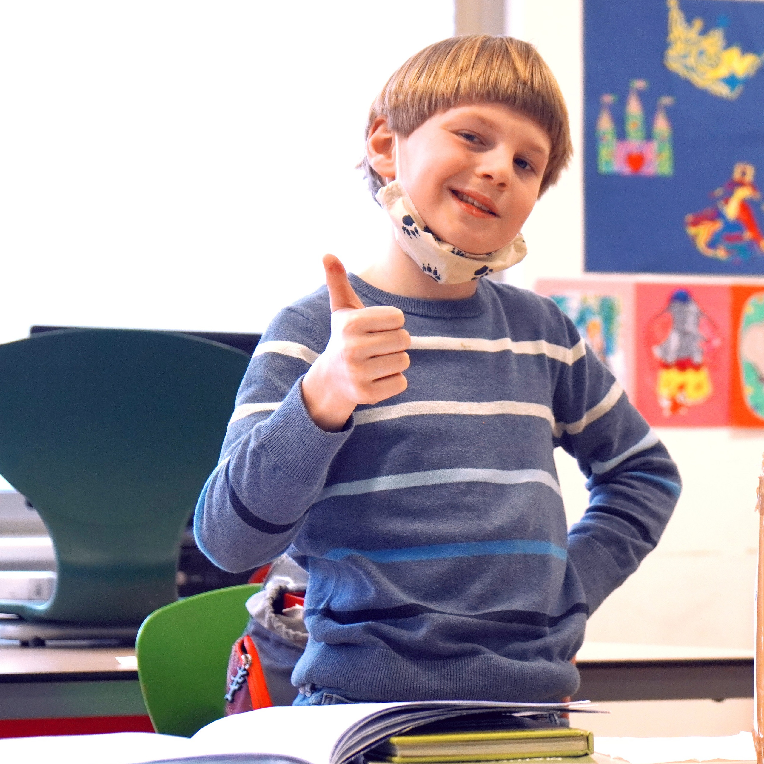 Ein junger Schüler zeigt mit dem Daumen nach oben und schaut stolz in die Kamera, neben ihm steht auf dem Schultisch eine selbstgebastelte Aktentasche aus Karton, auf der in großen Buchstaben Akten steht.