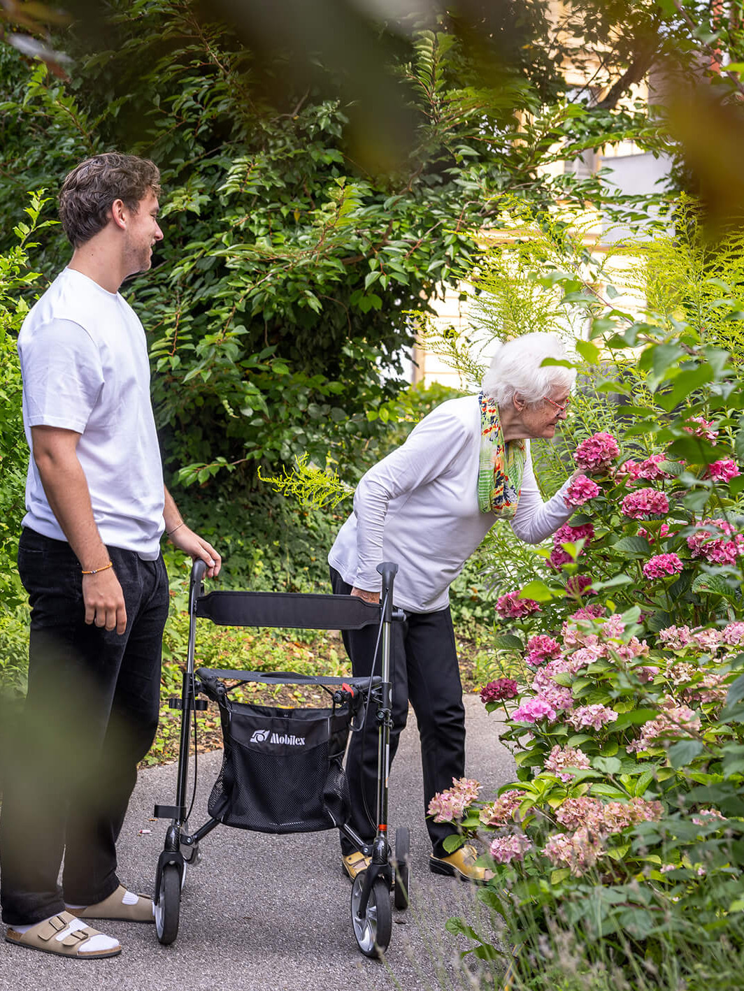 Ein Mann und eine Frau gehen gemeinsam durch einen Garten; die Frau beugt sich vor und riecht an einer Blume.