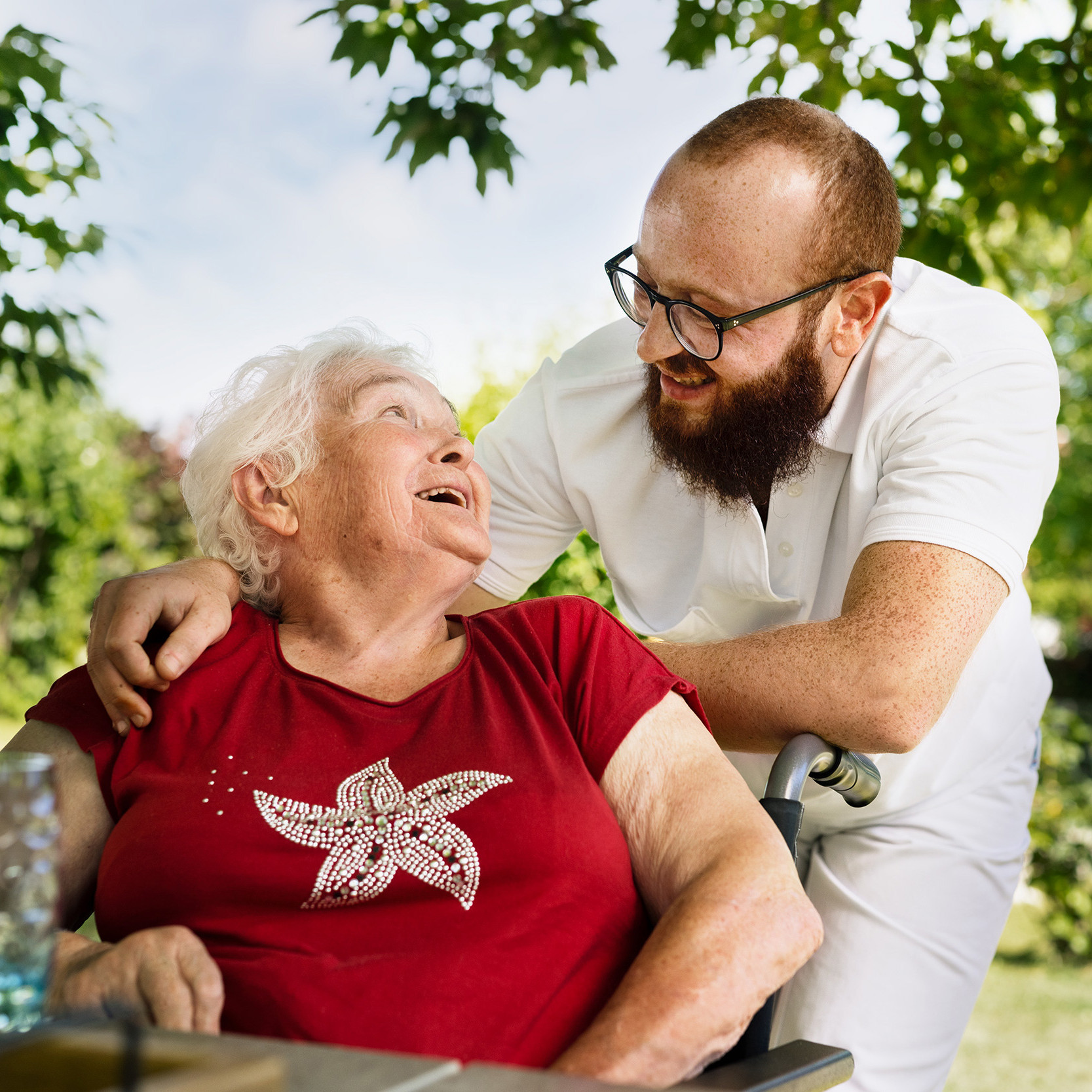 Ein Pfleger und eine ältere Damen im Rollstuhl lachen miteinander.