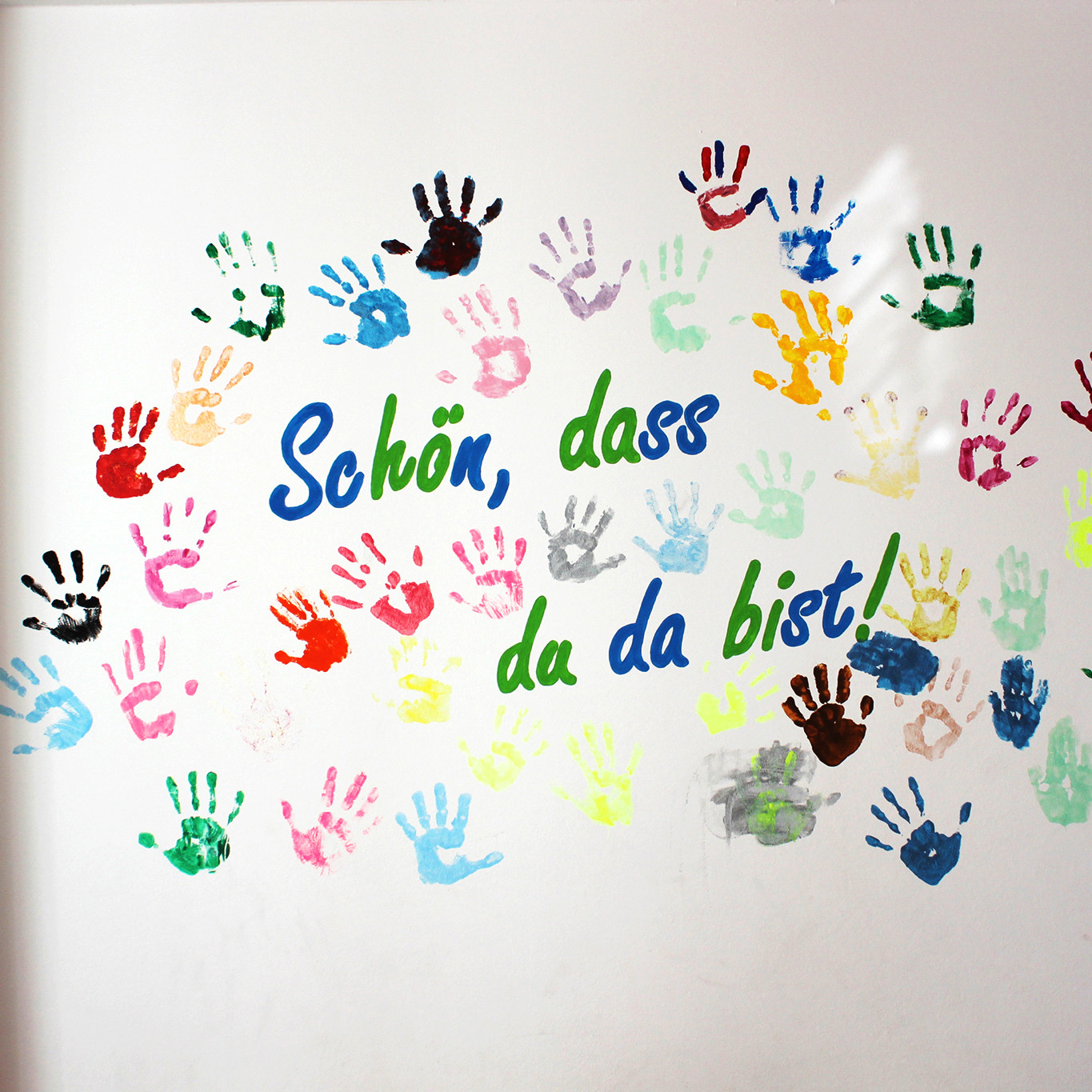 Viele bunte Farabdrücke von Kinderhände auf einer weißer Wand, dazu folgender Schriftzug: Schön, dass du da bist!