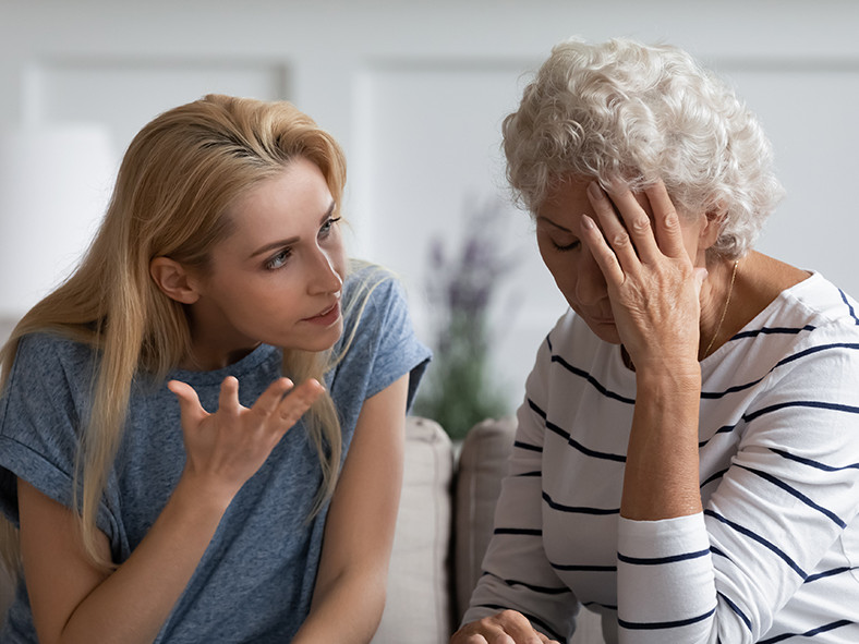 Eine jüngere Frau im Gespräch mit älterer Dame, die sich überfordert das Gesicht hält.
