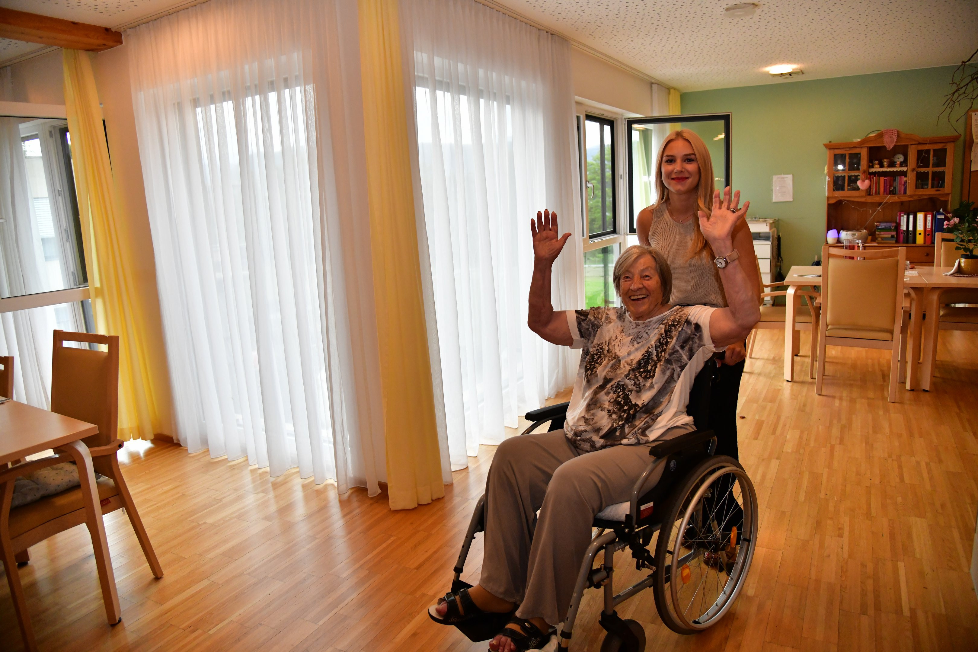 Eine ältere Person im Rollstuhl und eine junge Person dahinter, beide lachen