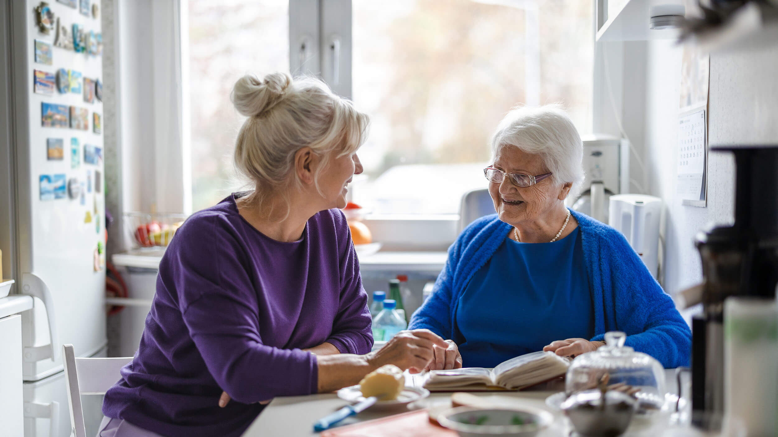 Zwei Frauen, eine davon älter, sitzen an einem Küchentisch und unterhalten sich.