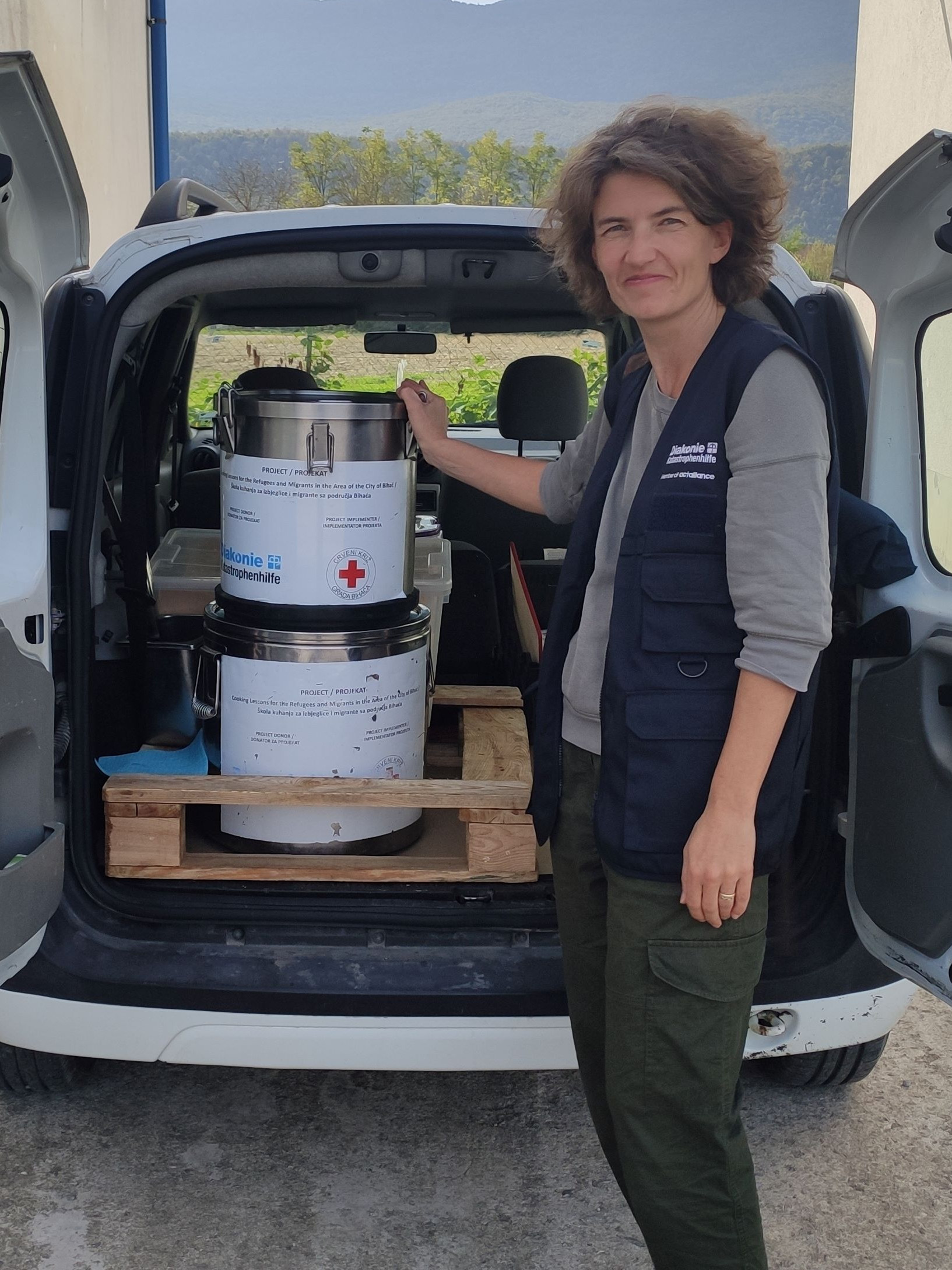 Mitarbeiterin der Diakonie Katastrophenhilfe steht vor einem Auto, das mit großen Essenstöpfen beladen ist.