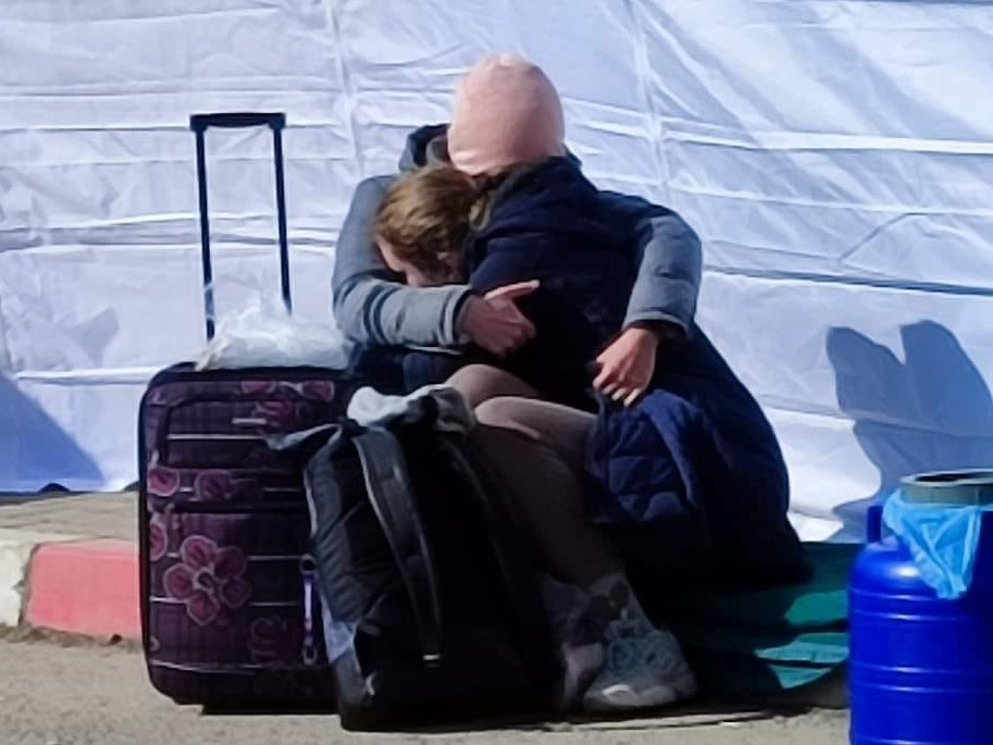 Eine Frau und ihre jugendliche Tochter sitzen mit Reisegepäck auf einem Gehsteig und umarmen sich.