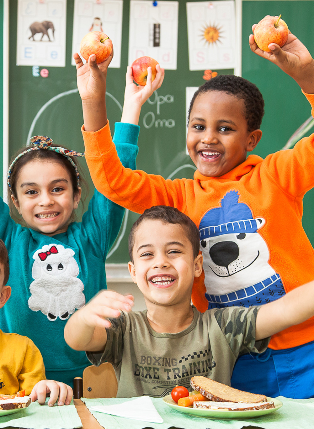Lachende Kinder vor einer Schultafel halten frische Äpfel in die Luft.
