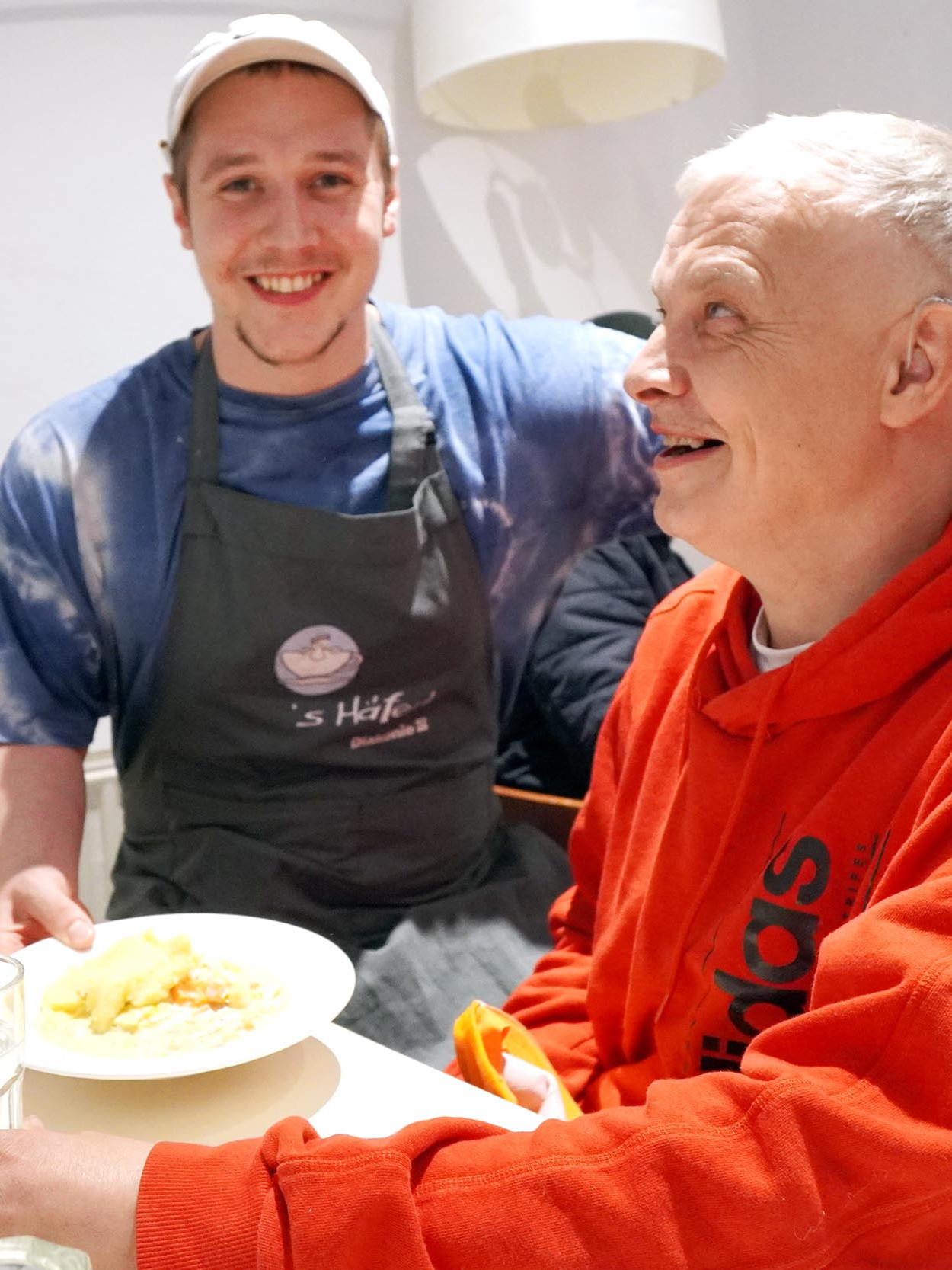 Ein junger Mann mit Schürze serviert einem Mann, der am Tisch sitzt, ein Essen.