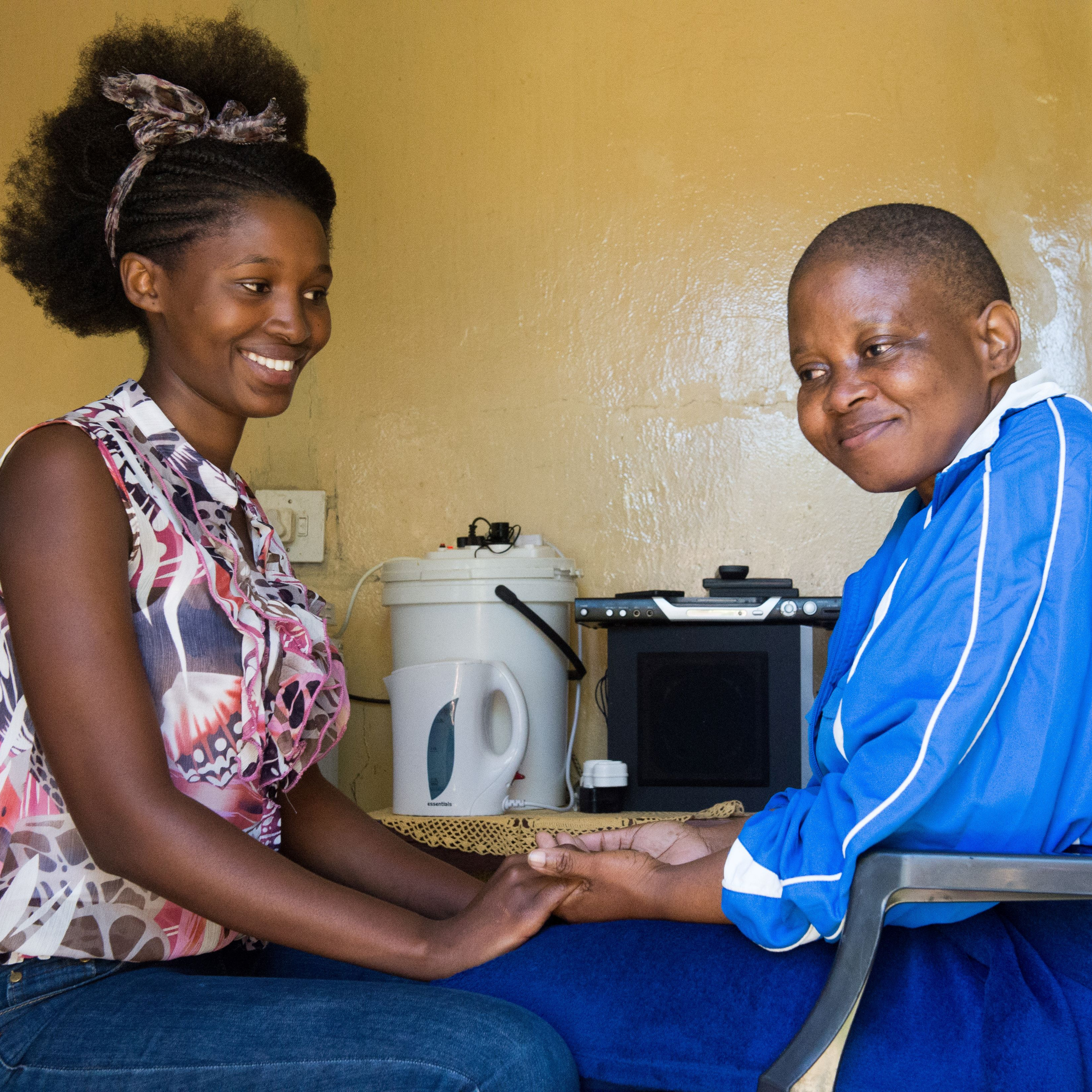 Zwei Frauen sitzen gegenüber, halten einander an beiden Händen und lächeln.