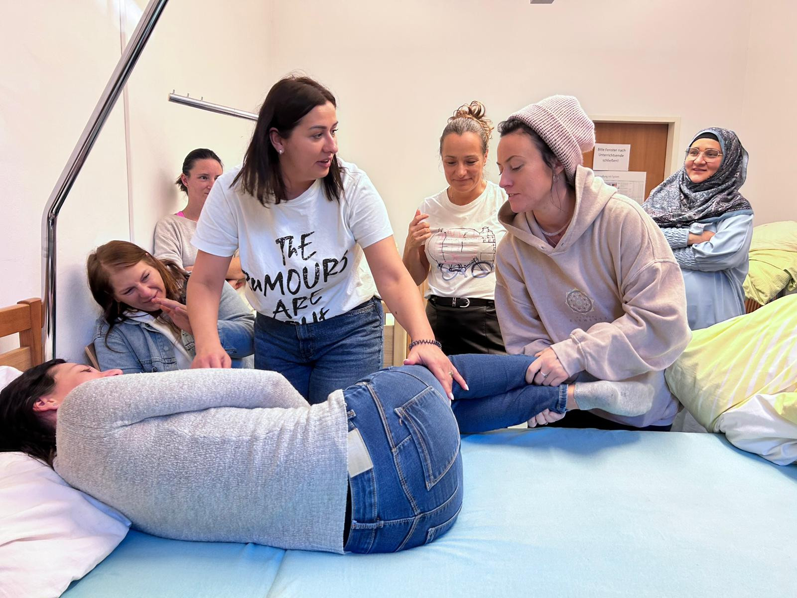 Eine Person liegt auf einem Bett und wird von zwei Personen richtig positioniert, weitere Personen sehen genau zu
