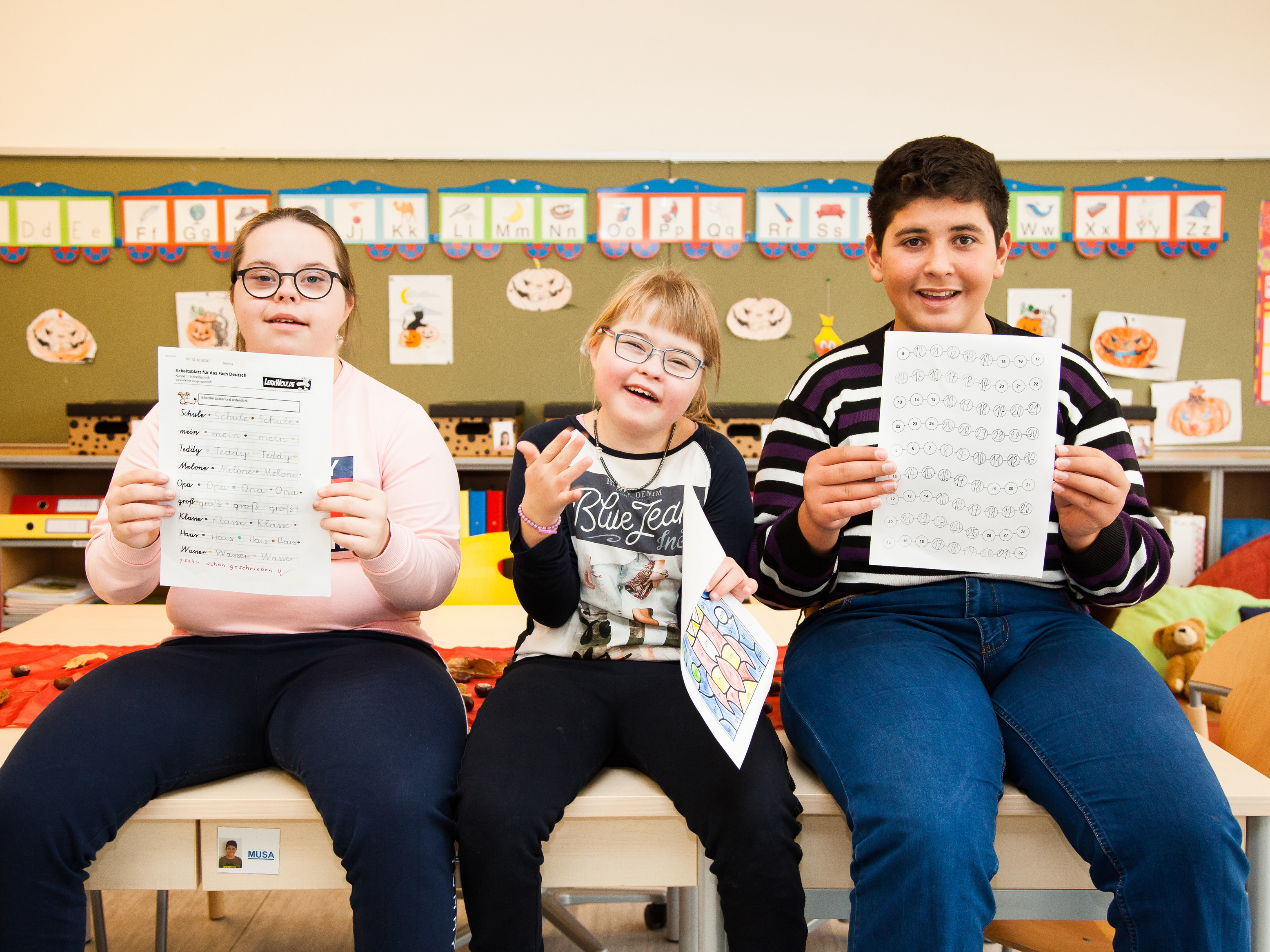 Drei Kinder mit und ohne Behinderungen lachen in die Kamera. Sie sitzen auf Tischen und halten Übungsblätter hoch.