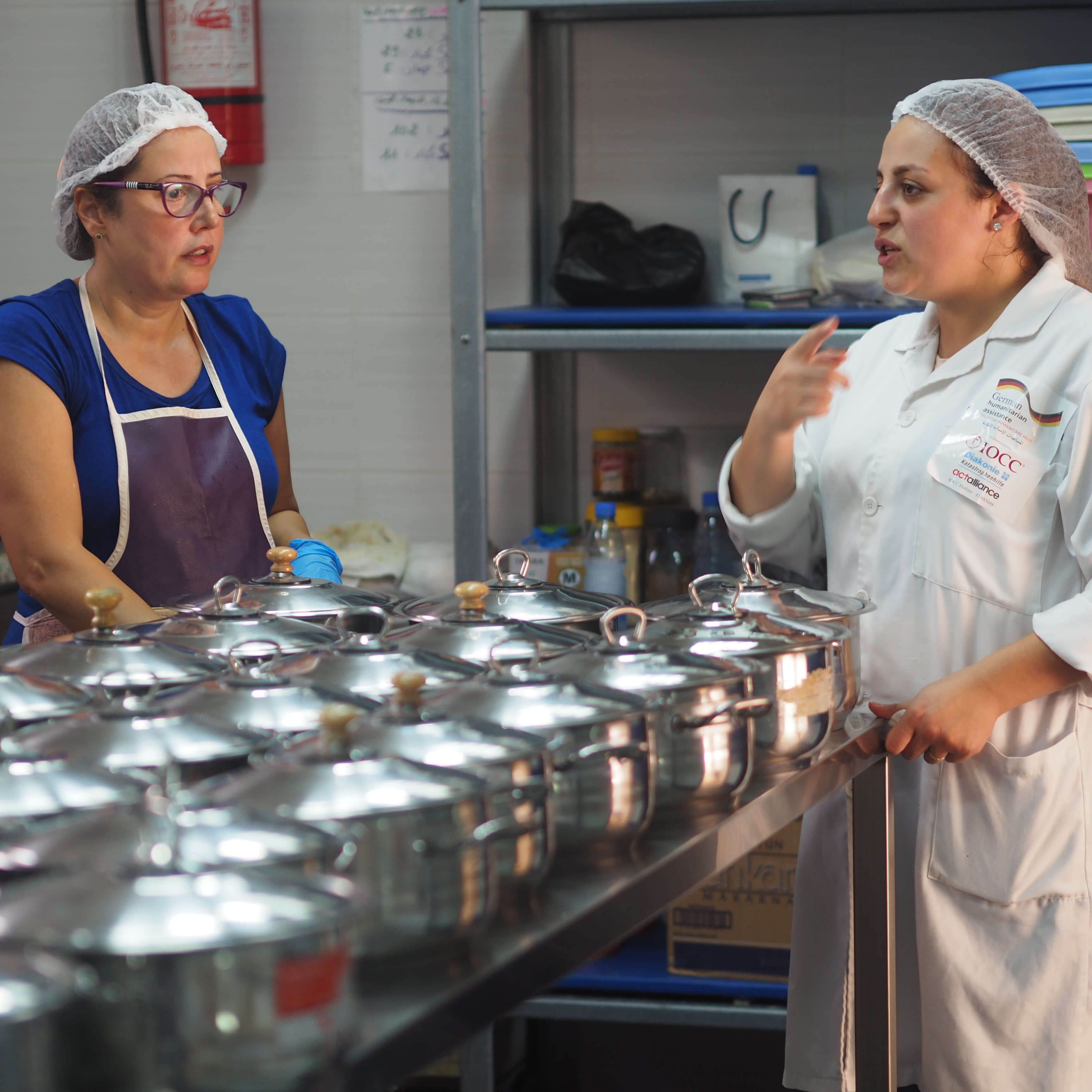 Zwei Köchinnen unterhalten sich, viele Kochtöpfe aus Metall stehen vor ihnen aufgereiht.