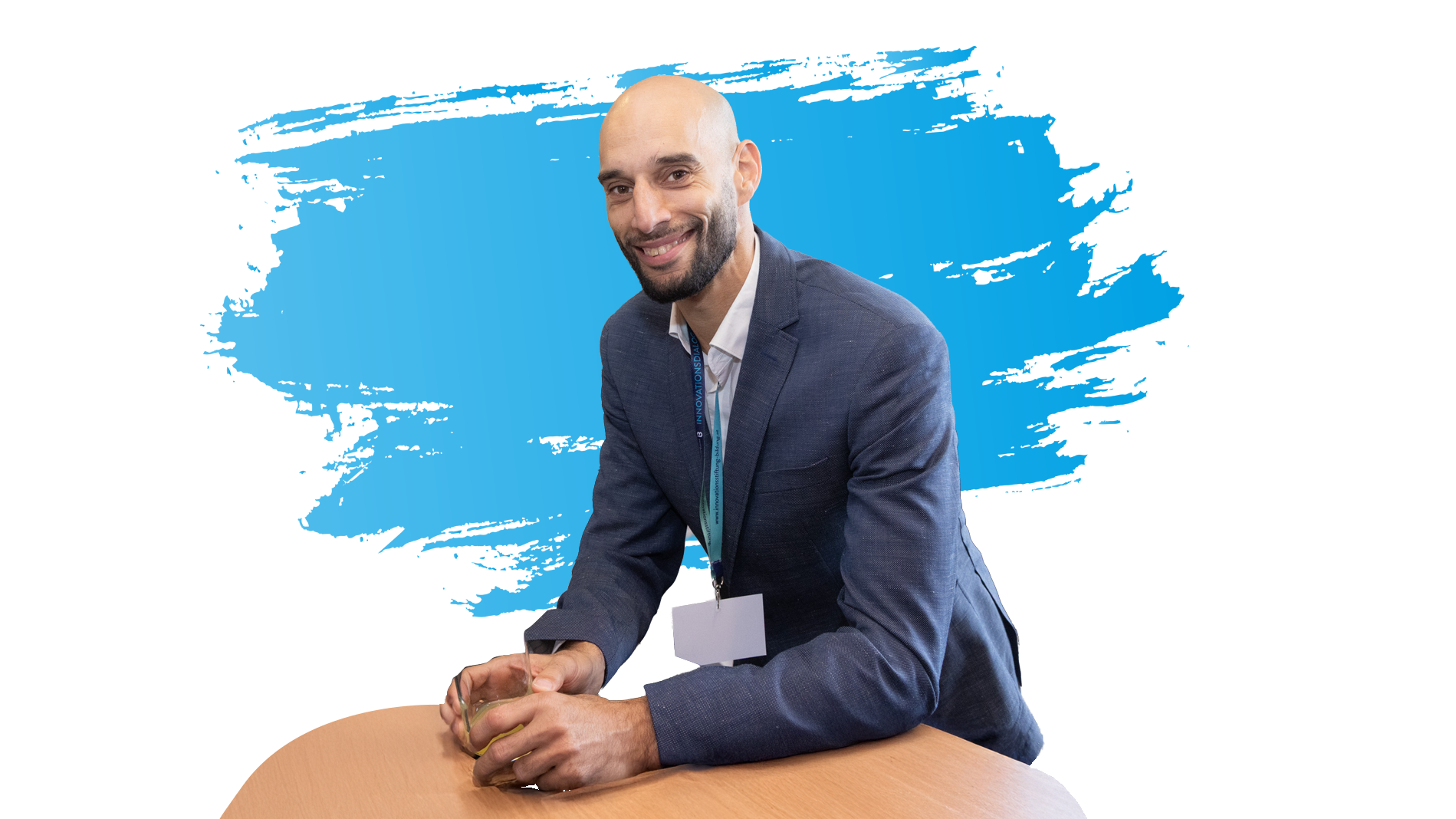 Ein Mann lehnt an einem Tisch und lächelt in die Kamera. Im Hintergrund ist ein blauer Farbstrich.
