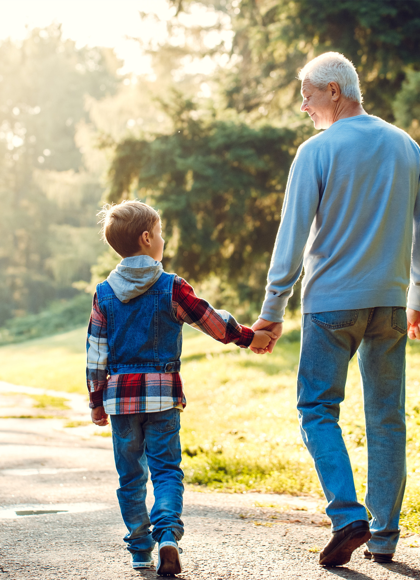 Ein Bub und ein älterer Mann gehen Hand in Hand einen Weg in der Sonne entlang.