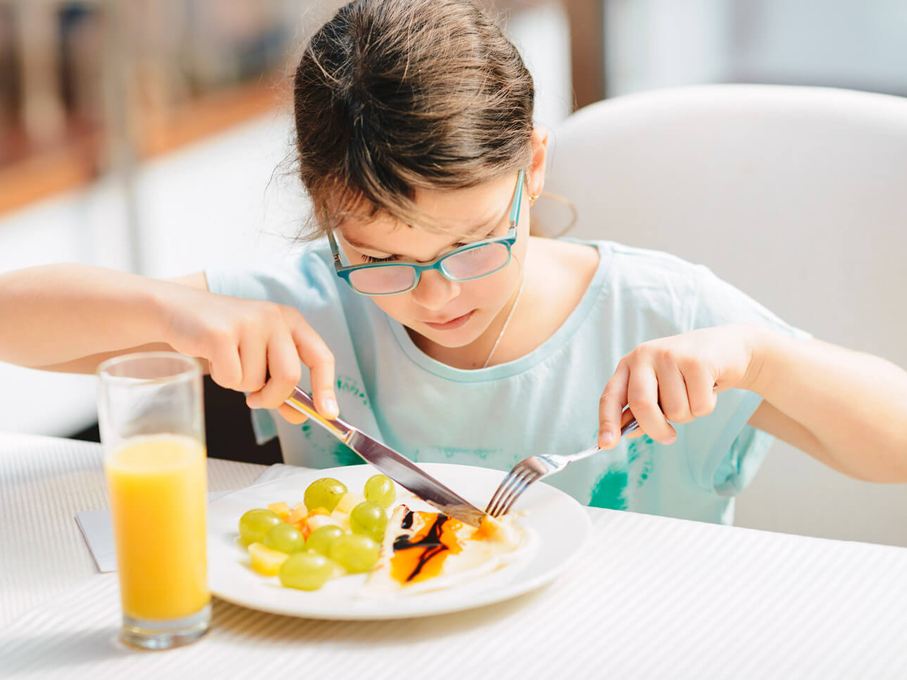 Ein Mädchen sitzt am Tisch und isst Obst und Gemüse mit Gabel und Messer. Daneben steht ein Glas Orangensaft. Ihr Blick ist auf den Teller gerichtet.