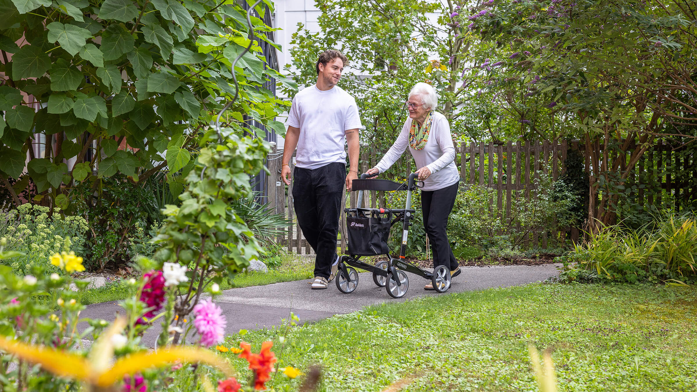 Ein Person mit Rollator und ein Pfleger unterhalten sich während eines Spazierganges im Garten