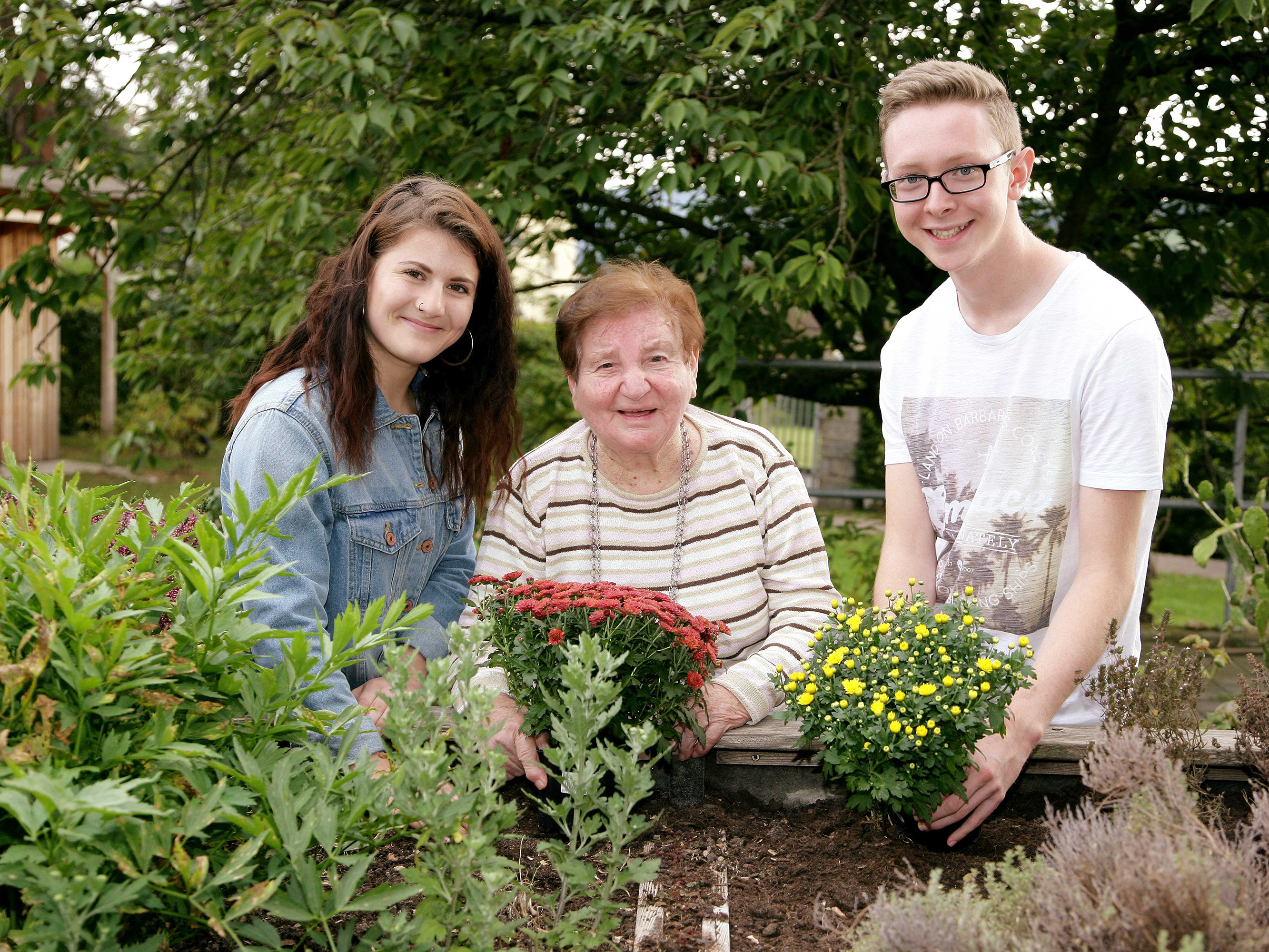 Zwei junge Menschen und eine Frau im Alter setzen gemeinsam Blumen in ein Hochbeet.