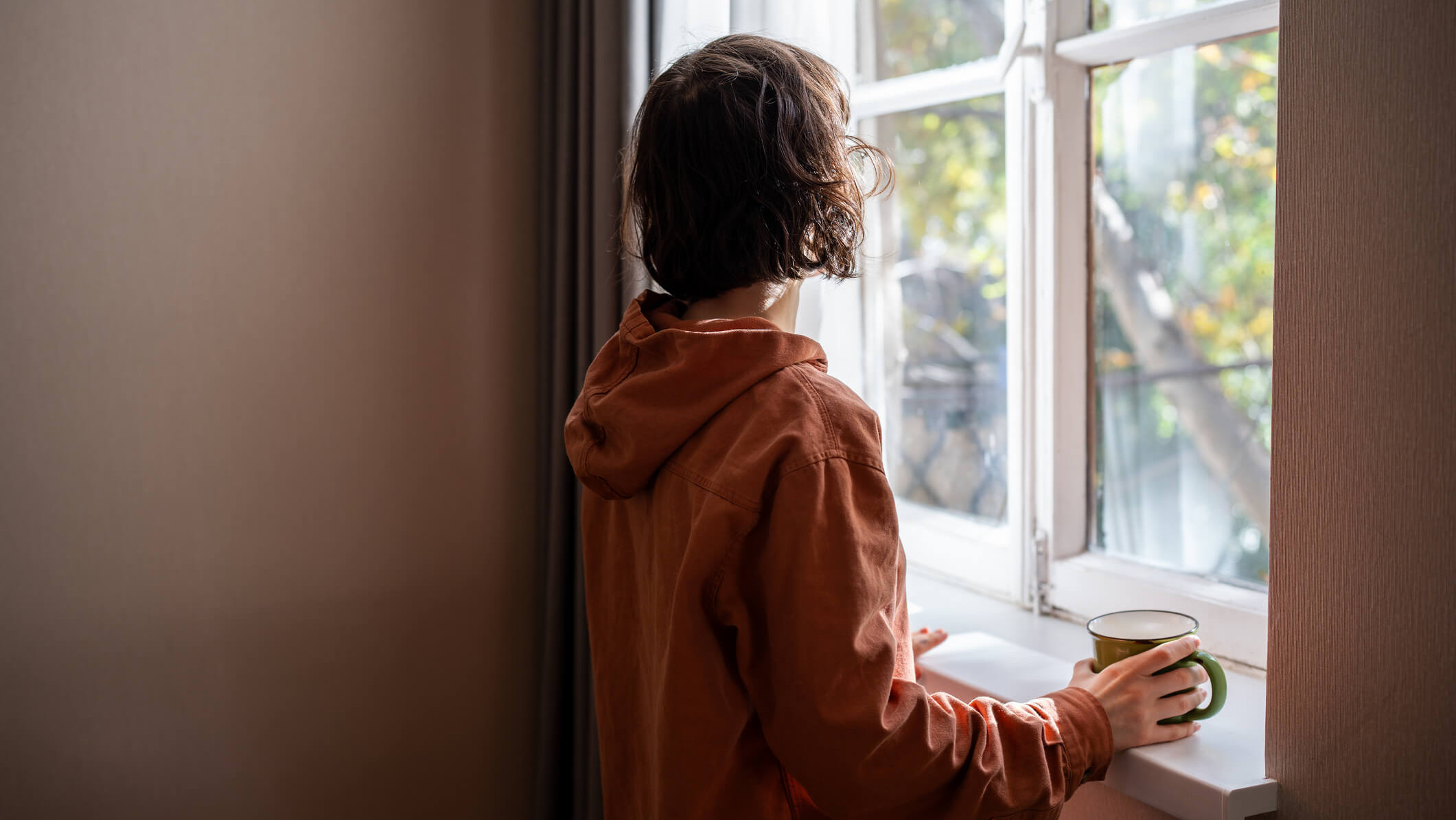 Die Frau steht am Fenster und blickt hinaus. In der Hand hält sie eine Tasse.