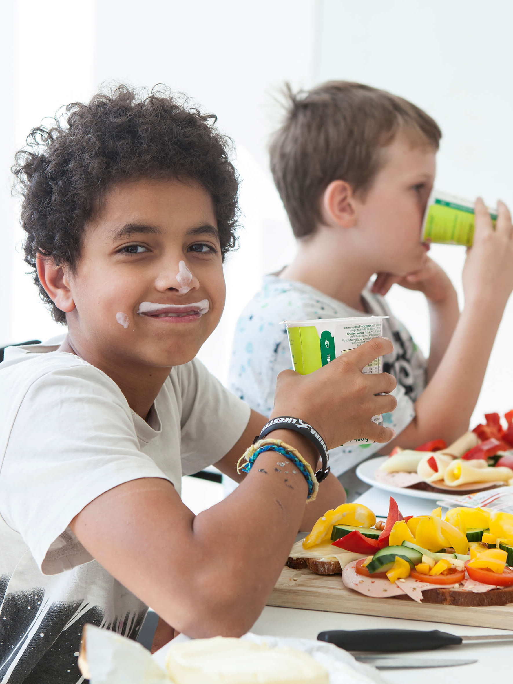 Kinder sitzen am Tisch und genießen eine gesunde Jause mit Apfel, belegten Broten und Gemüse. Zwei Buben trinken Joghurt – einer ist bereits fertig und hat Joghurt an Nase und Mund.