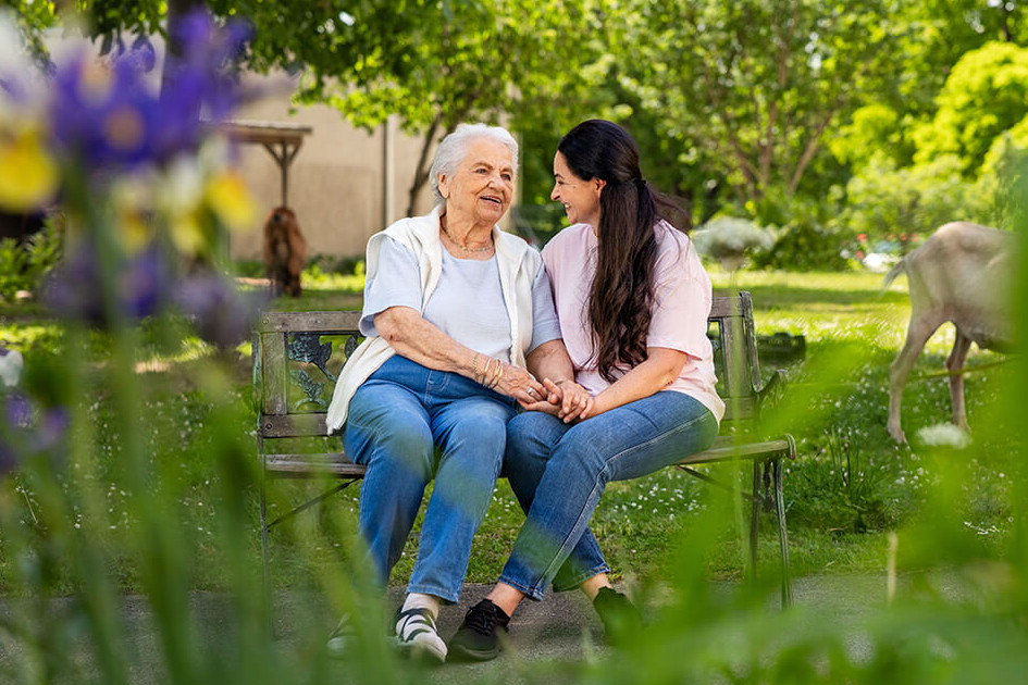 Eine ältere und eine jüngere Frau sitzen lächelnd auf einer Parkbank in einem Garten. Im Hintergrund ist eine Ziege zu sehen.