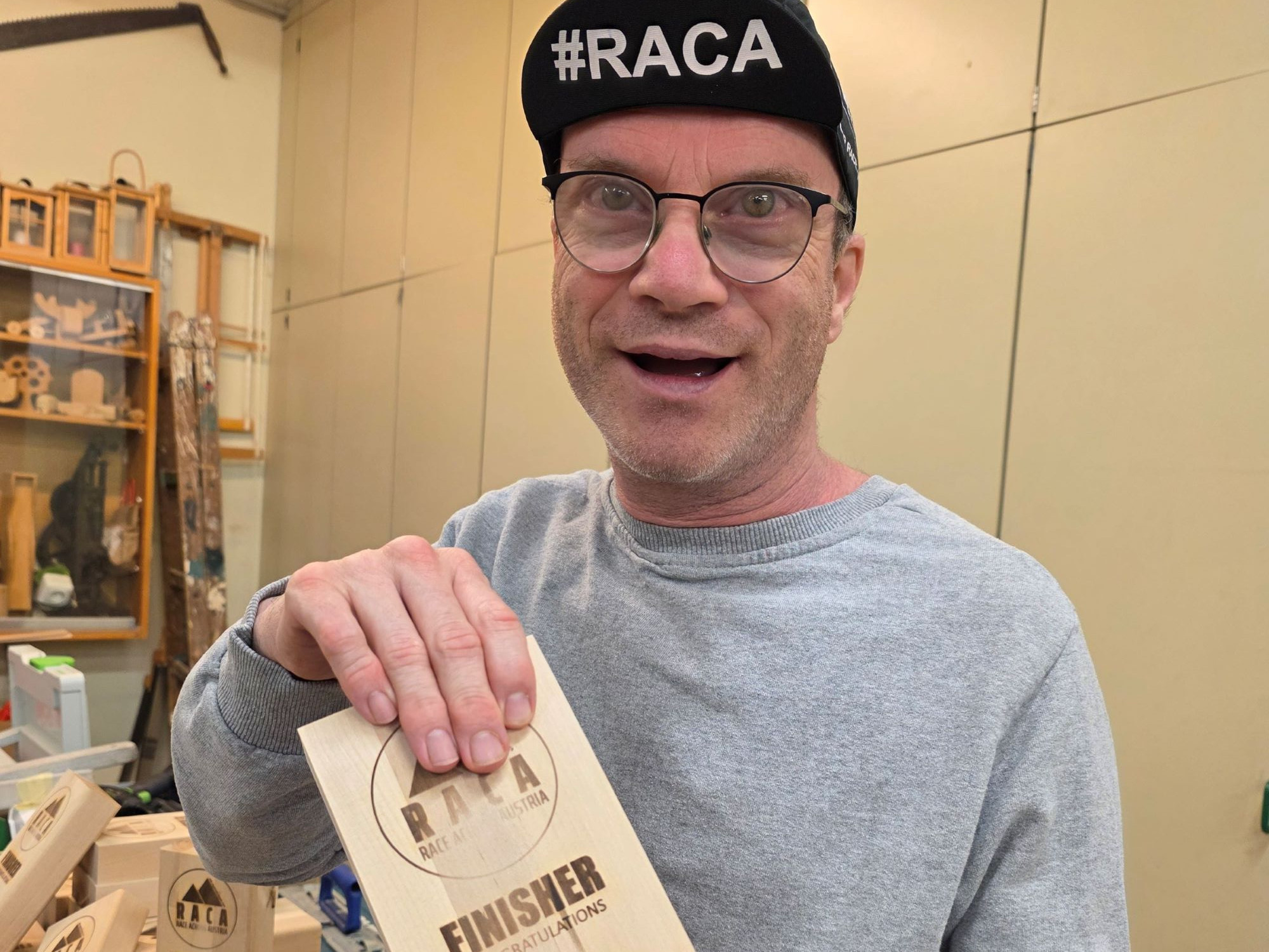 Mann in hellgrauem Pullover, Brille und schwarzer Kappe hält Holztrophäe in Händen
