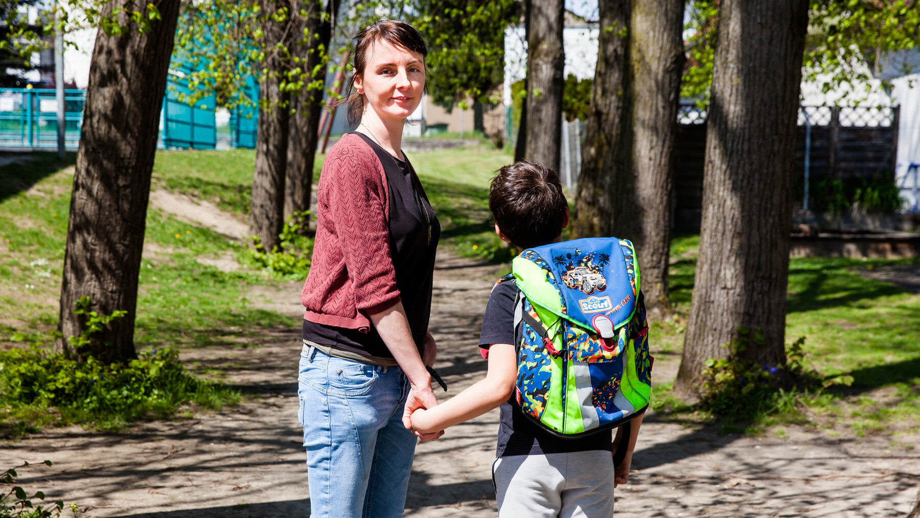 Eine junge Frau und ein Bub halten einander an der Hand. Der Bub hat eine Schultasche am Rücken, er ist von hinten zu sehen. Die Frau schaut in die Kamera.