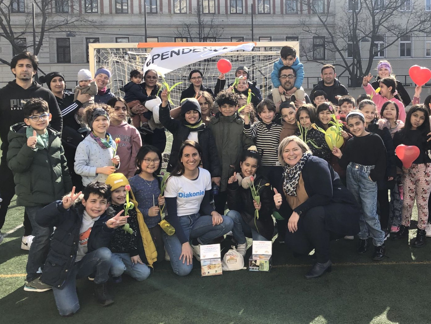 Volksschulkinder mit Eltern und Lehrer:innen auf einem Sportplatz vor der Schule. Einige halten rote herzförmige Luftballons
